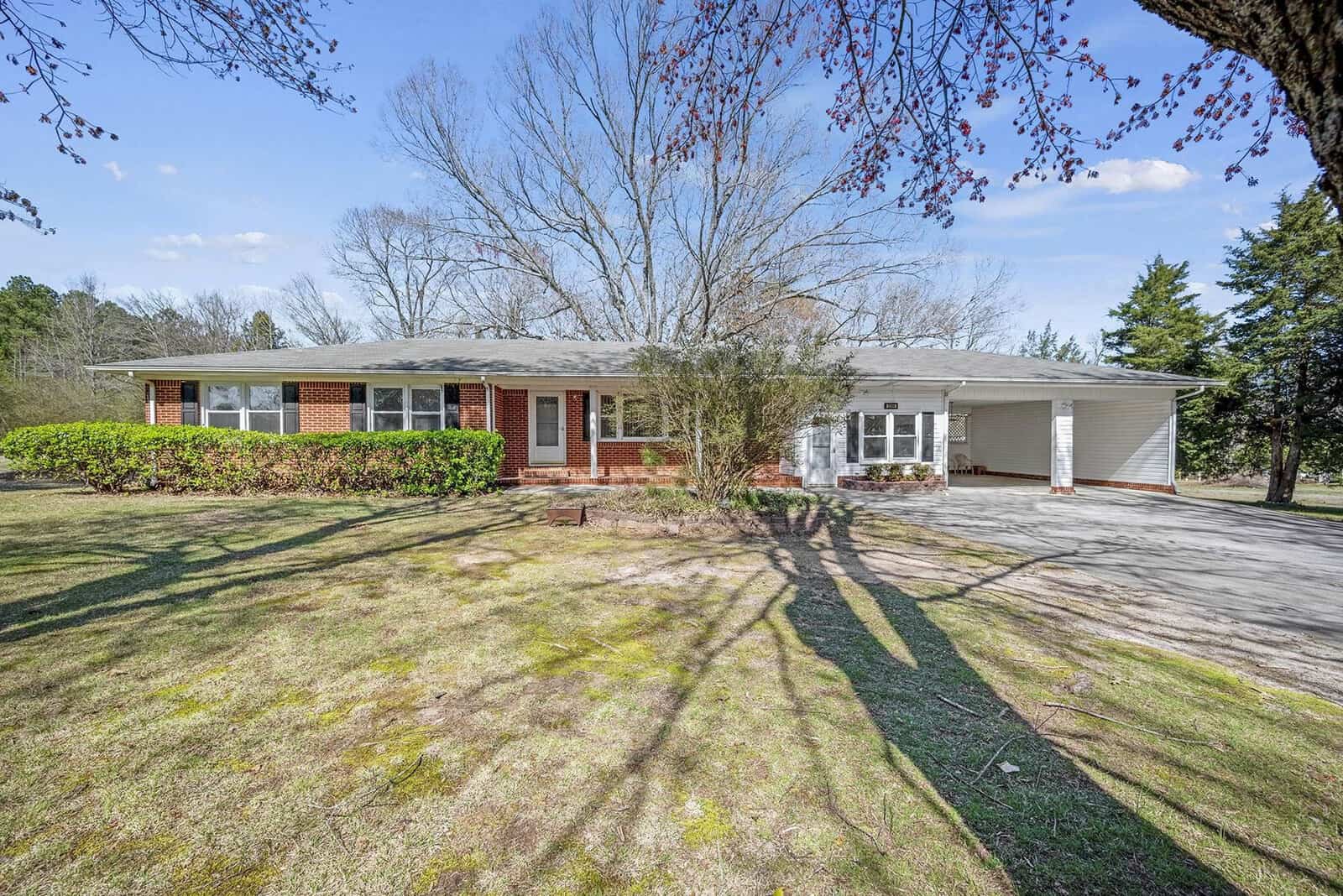 Single-story brick house with an attached carport, situated in a sunny, grassy area with trees casting shadows in the yard.