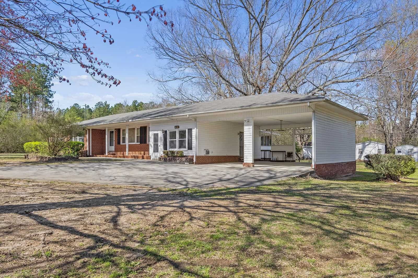 A single-story house with a carport on a sunny day, featuring a brick foundation and a front porch with white columns, surrounded by a lawn with scattered trees and shadows.