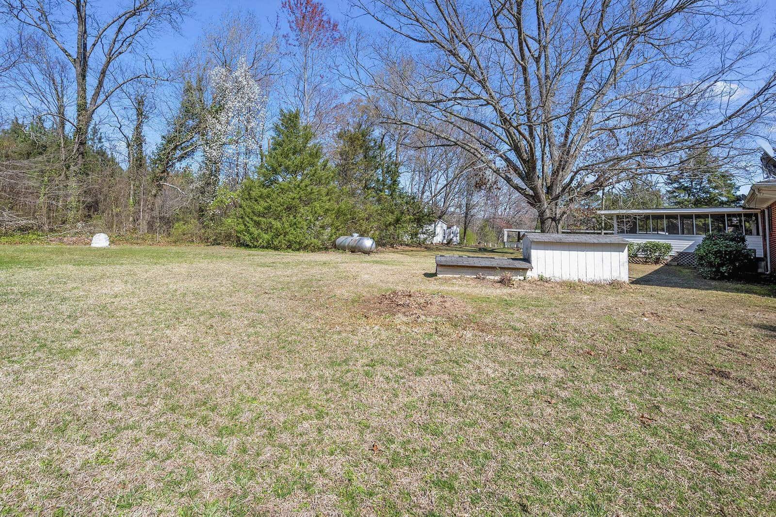A spacious backyard with a well-maintained lawn, featuring a large propane tank to the left, a small wooden bench in the center, and a white railing leading to a house's deck on the right, surrounded by leafless trees suggesting a late autumn or winter season.