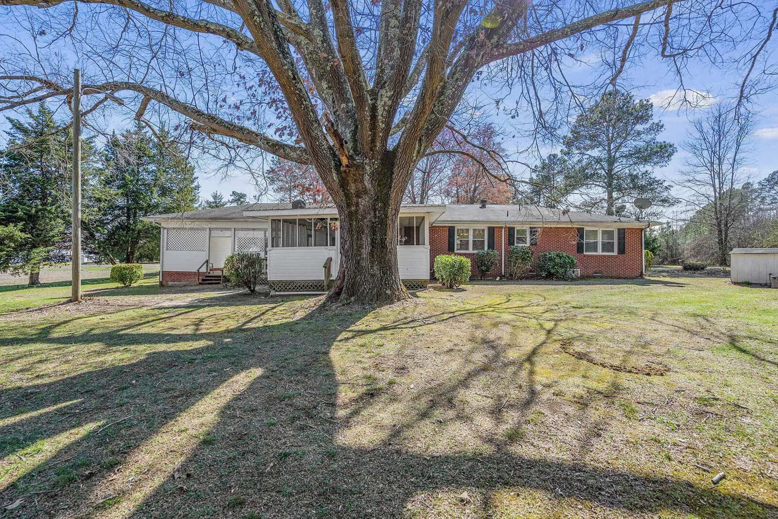 Single-story red brick house with white siding, featuring a front porch and large tree in the yard, under a clear sky.