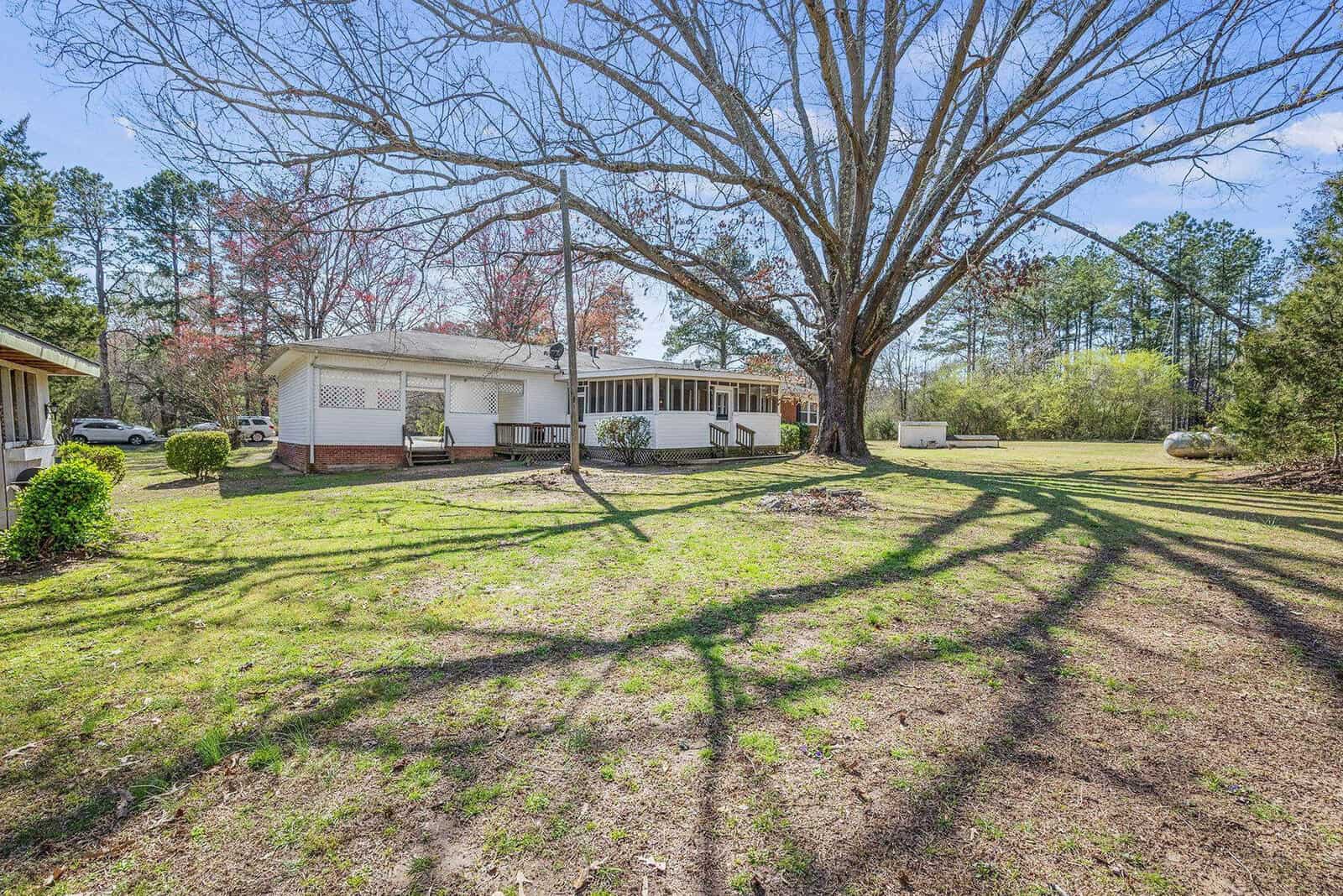 A single-story home with white siding under the expansive branches of a large tree on a sunny day, casting shadows on the green lawn.