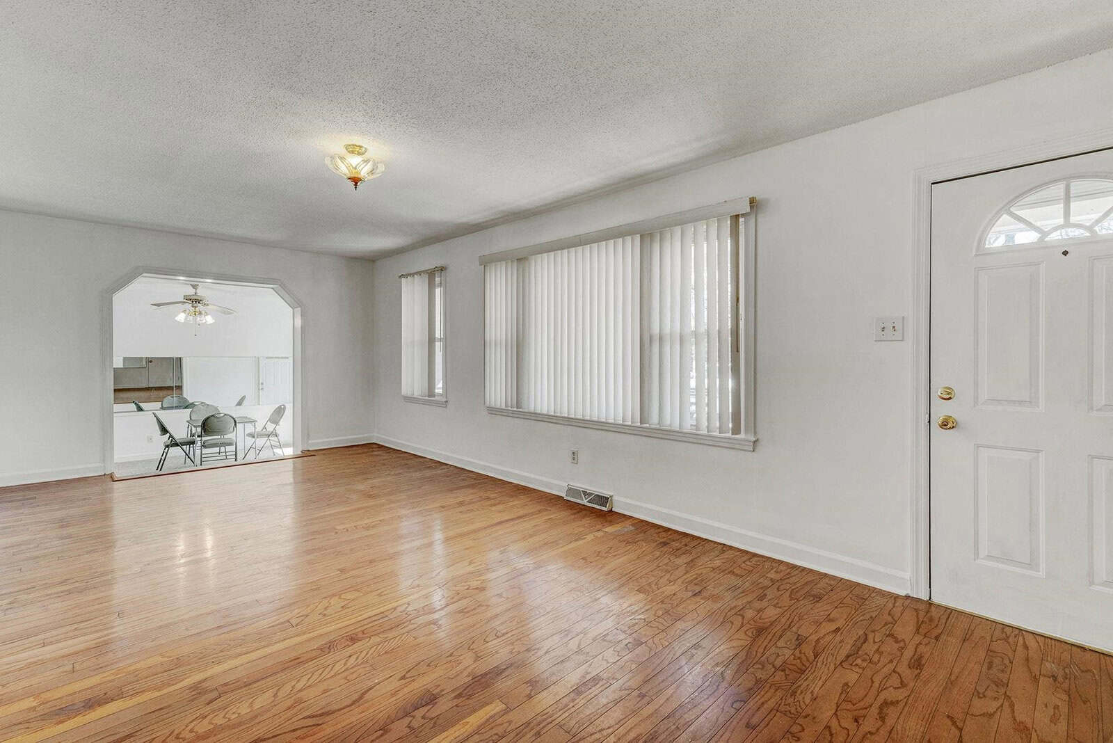 A spacious, empty room with hardwood floors, a white door, and a large window with vertical blinds. there is an arched doorway leading to another room with a dining table visible in the background.
