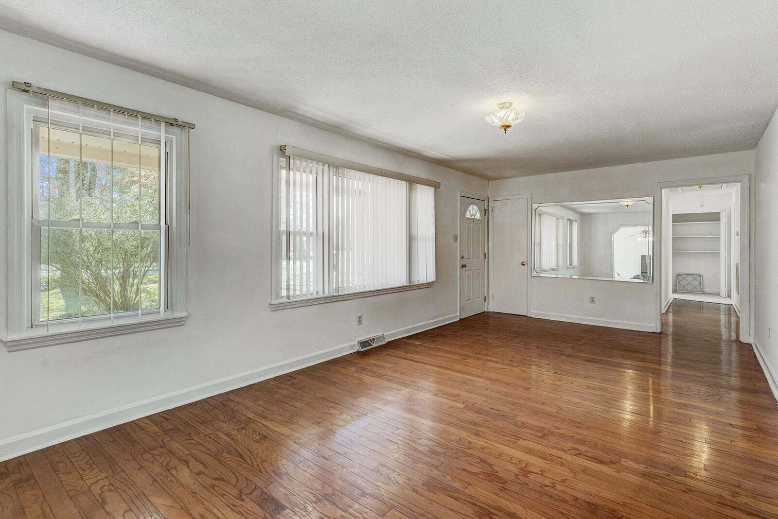 Empty living room with hardwood floors, white walls, and a ceiling light fixture, featuring two windows with blinds and an open doorway leading to another room.