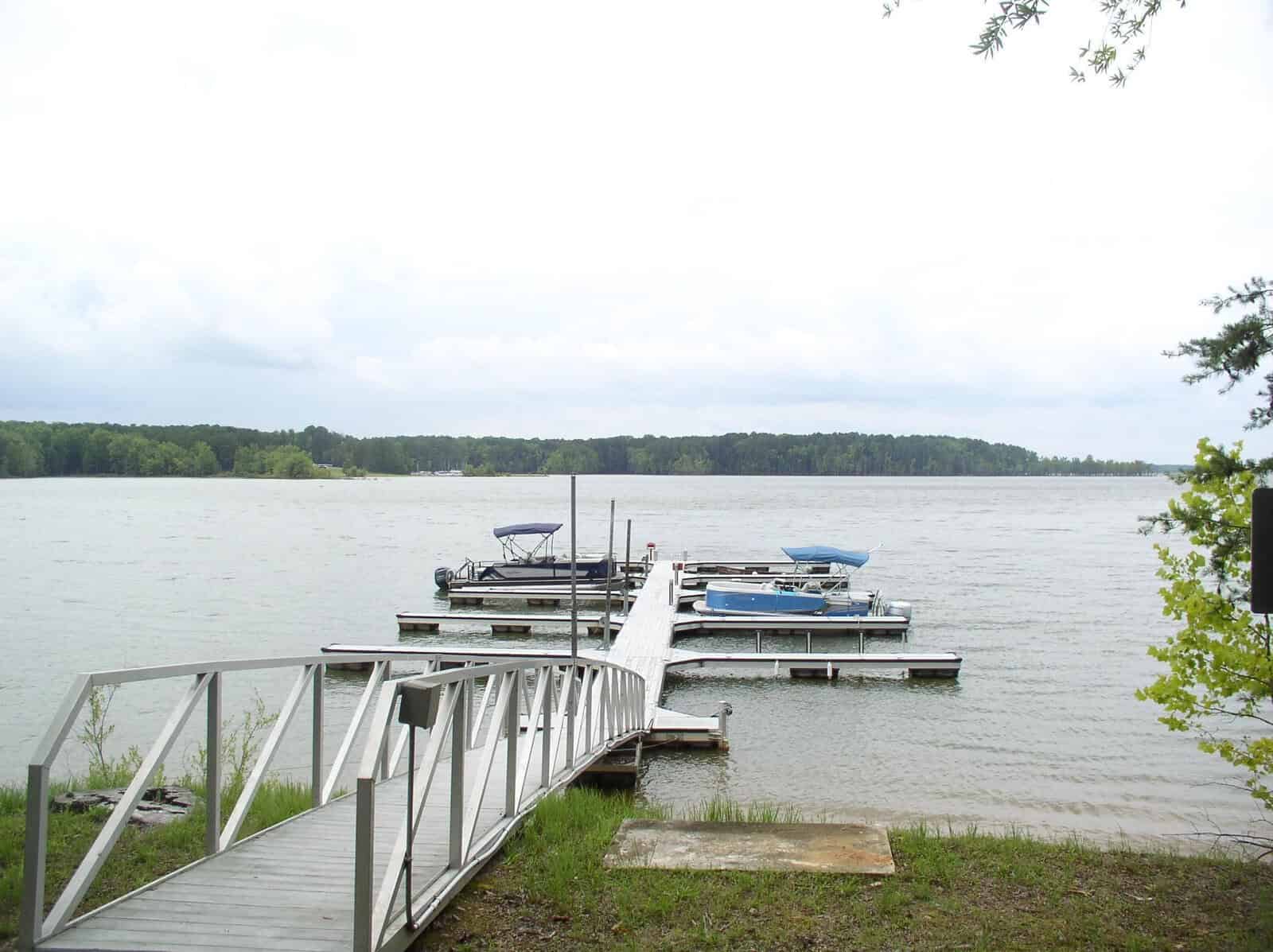 A metal walkway at 110 Somerset Lane, Henderson NC, leads to a small dock with several boats on a calm lake, surrounded by a forested area.
