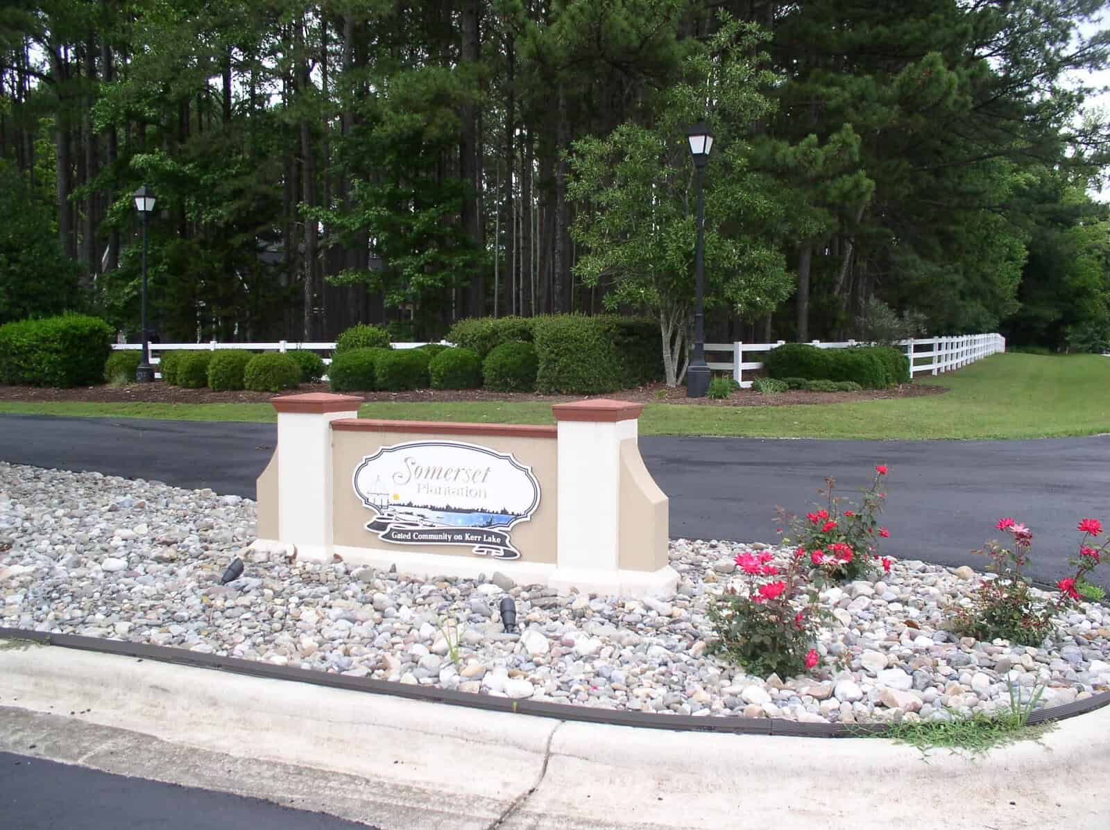 A stone sign with the inscription "somerset" stands at the entrance of a community, flanked by red flowers and surrounded by a landscaped area with white fencing in the background.
