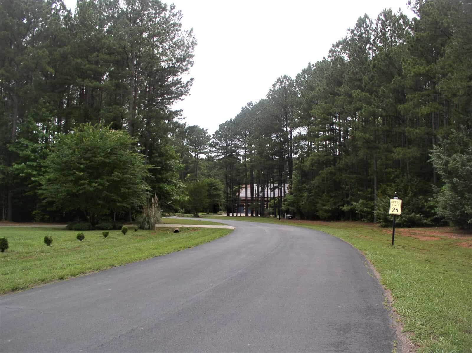 A winding road leading through a wooded area with lush green trees and a speed limit sign indicating 25 mph to the right.