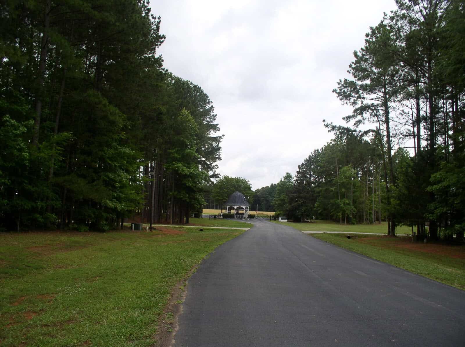A paved road leading towards a building with a blue roof, flanked by green trees and grass under a cloudy sky.