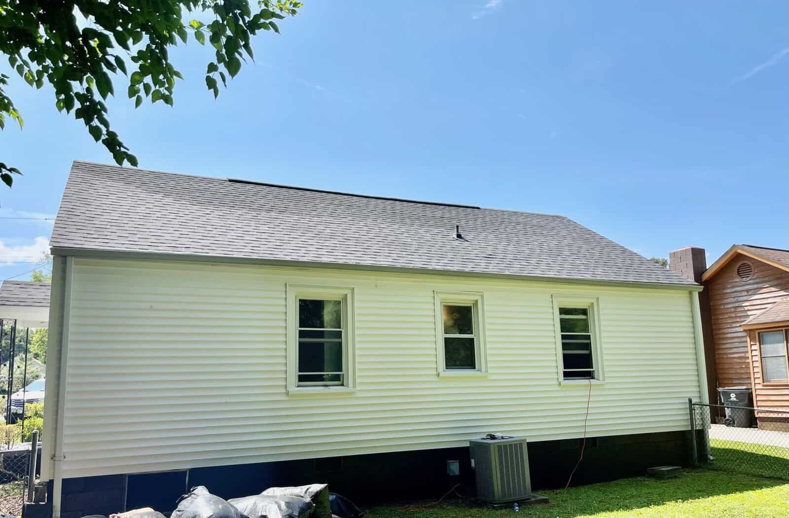 A single-story house with beige siding and a gray shingle roof under a blue sky with sparse clouds. the house has four windows and an air conditioning unit outside.