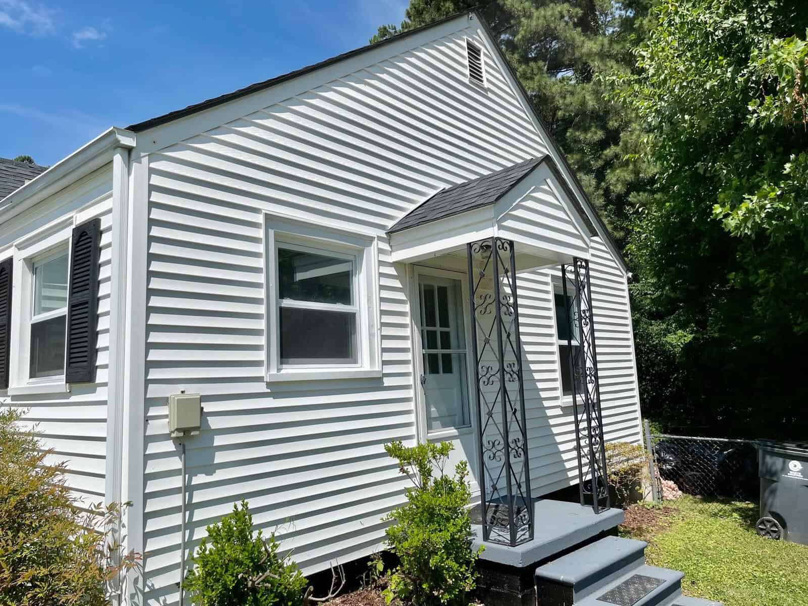 A small white house with vinyl siding, black shutters, and an ornate black metal screen door on a sunny day.