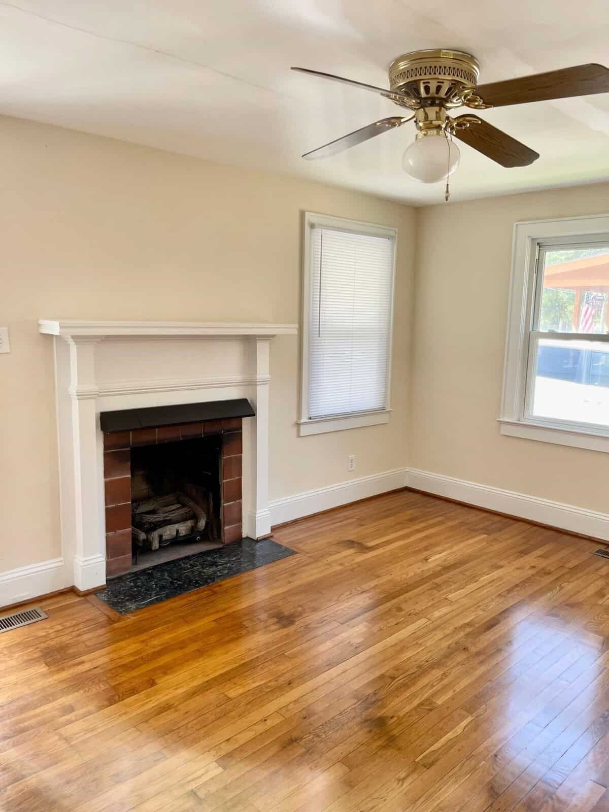 An empty room with polished wooden floors, a white fireplace, a ceiling fan, and two windows with white blinds.