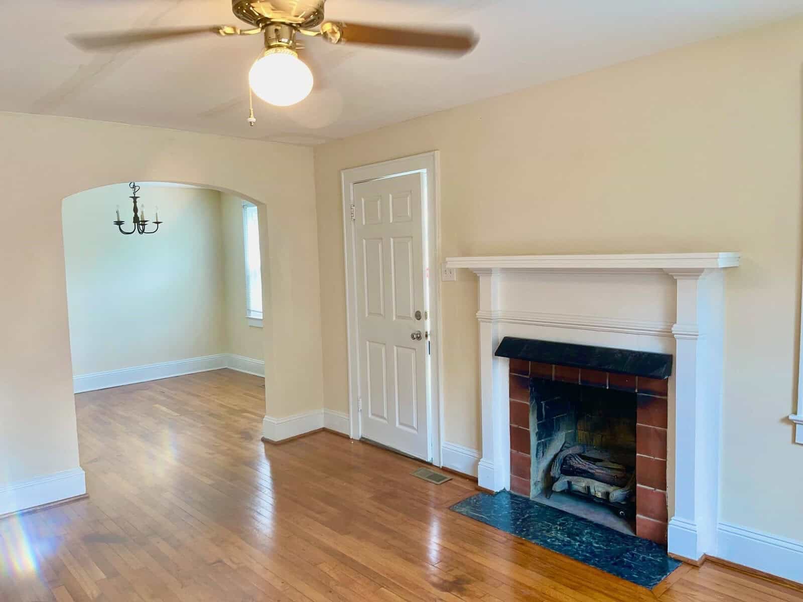 A well-lit, empty room featuring hardwood floors, a white fireplace, a ceiling fan with a light fixture, and a doorway leading to another room.