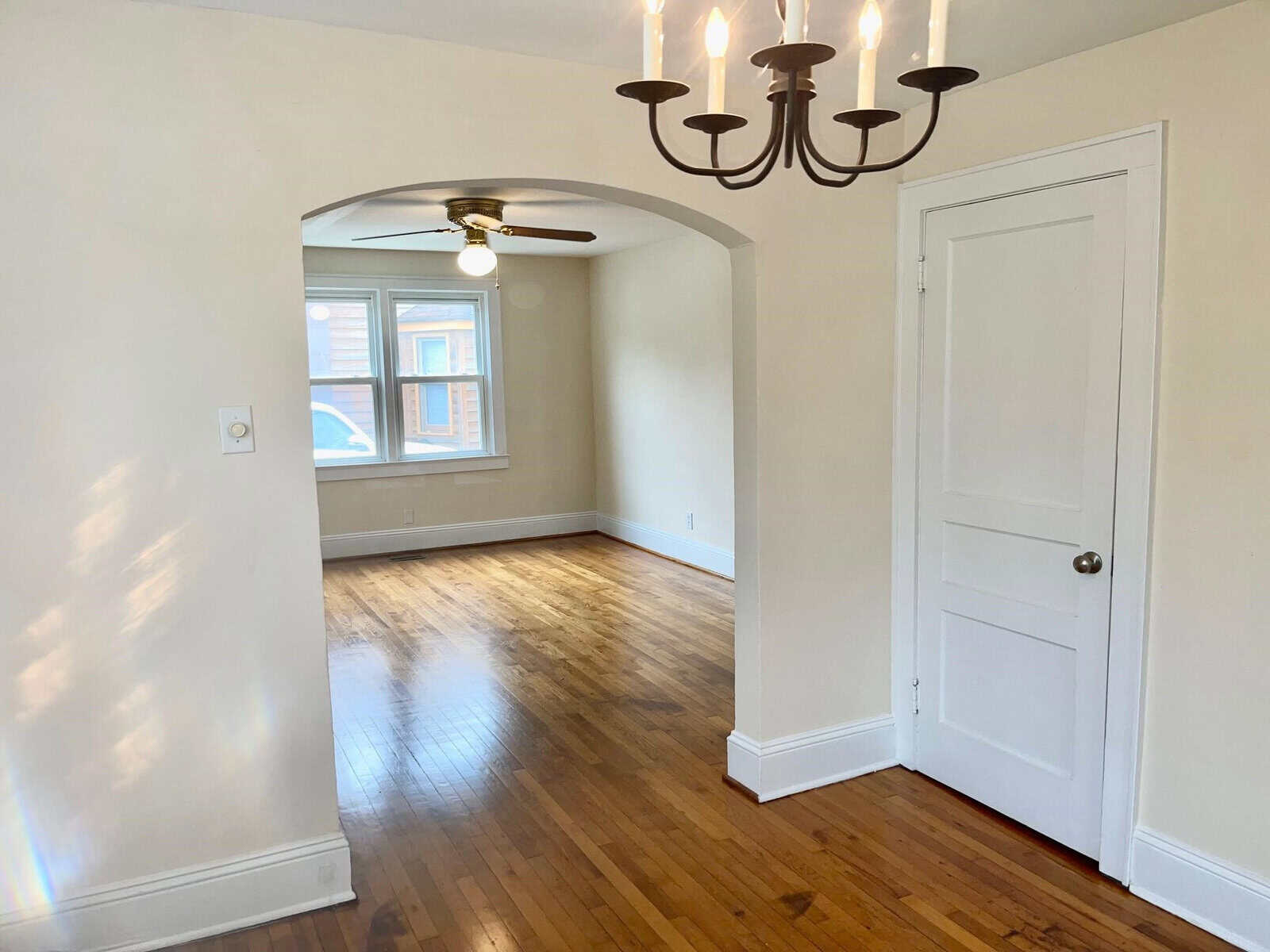 An empty room featuring polished hardwood floors, a white chandelier, and an arched entryway next to a closed white door, with natural light coming through windows.