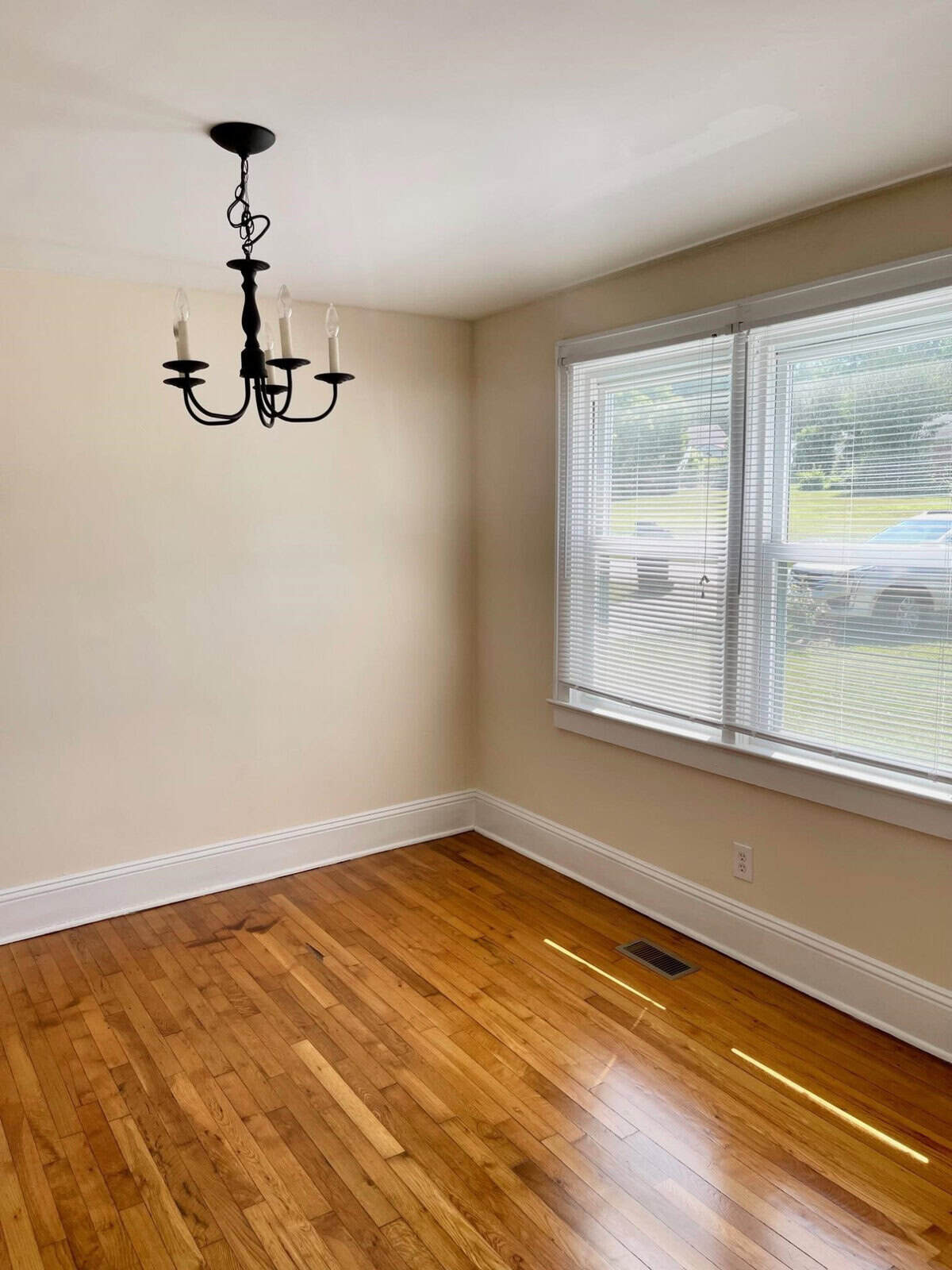 An empty room with beige walls, hardwood flooring, and a black chandelier, featuring a window with a view of an outdoor area.