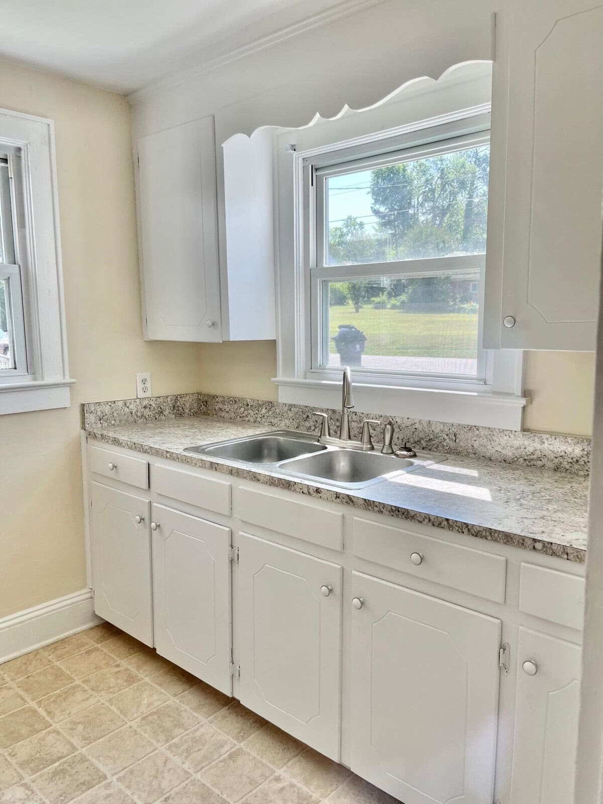 A bright, clean kitchen with white cabinetry, a stainless steel sink, and granite countertops, with a scalloped valance over a window that overlooks a grassy outdoor area.