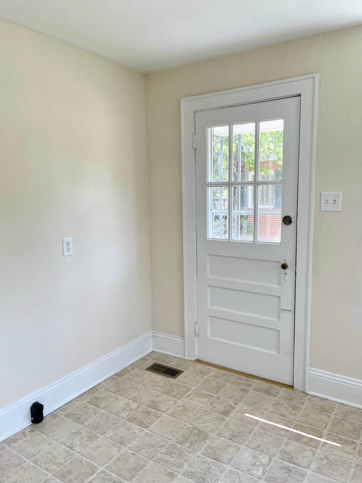 A sparsely furnished room with beige walls and tiled flooring, featuring a white door with a window and a black doorstop on the floor.