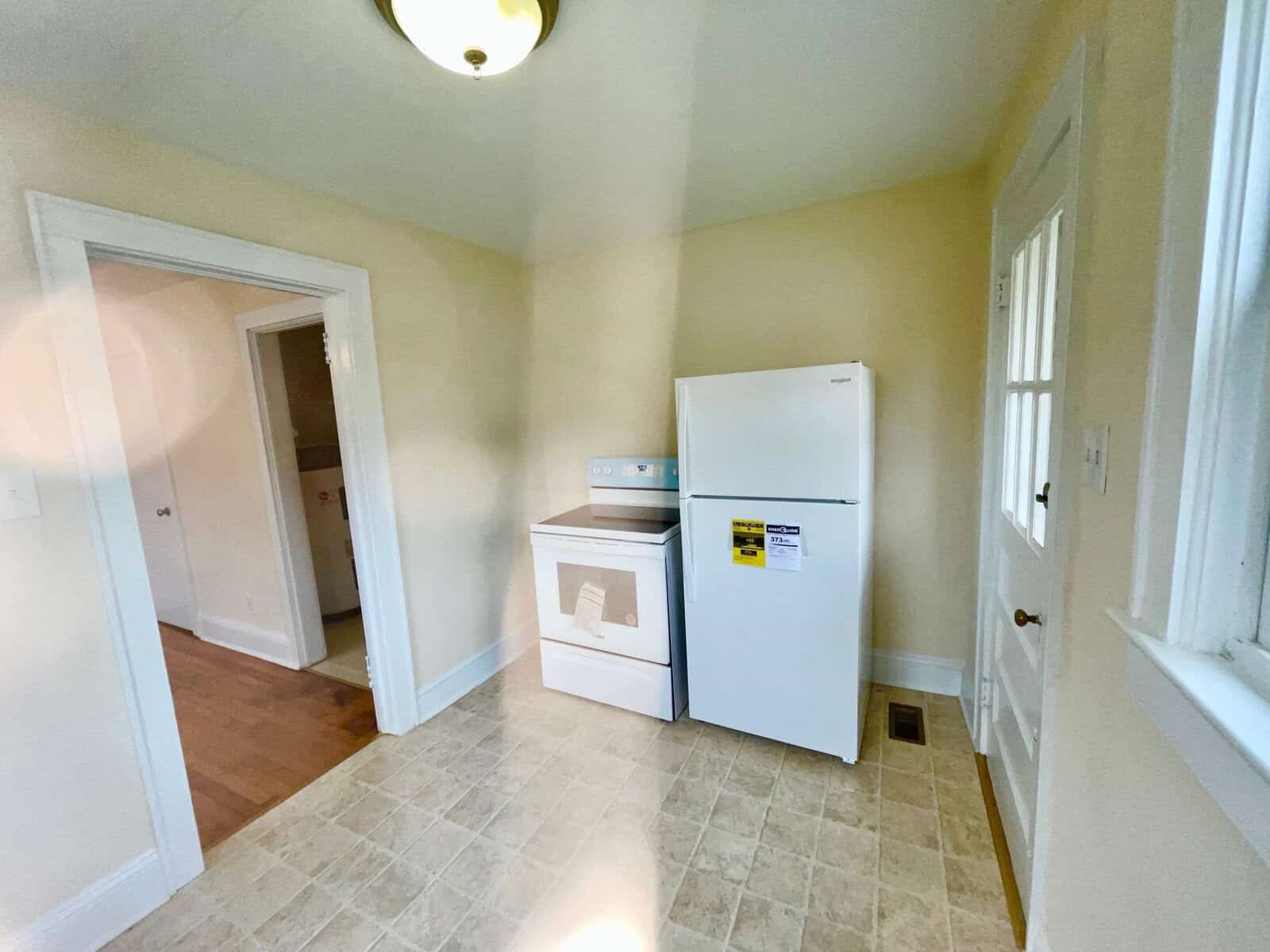 A small, empty kitchen area with white appliances, including a refrigerator and a stove, a tan tiled floor, and white walls illuminated by a ceiling light. there are three doors, one open, leading to other rooms.