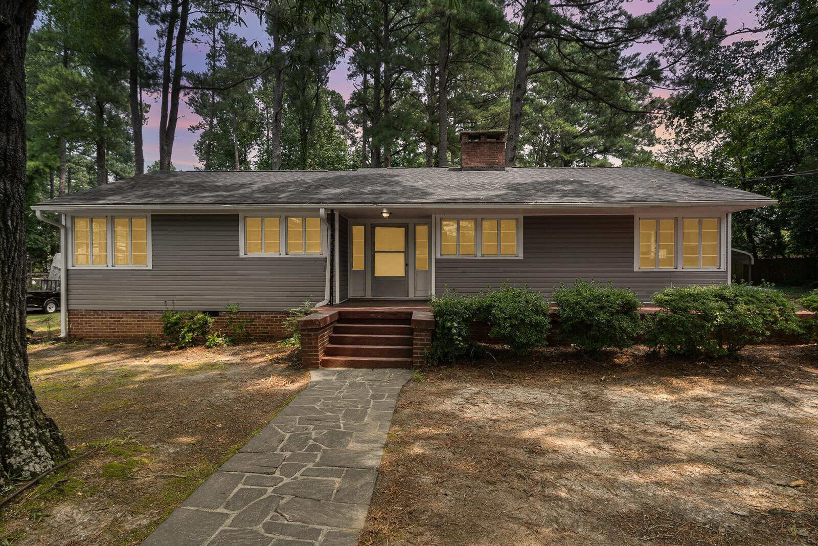 A single-story gray house with a chimney and yellow front door, featuring a stone pathway leading up to the entrance and surrounded by tall pine trees.
