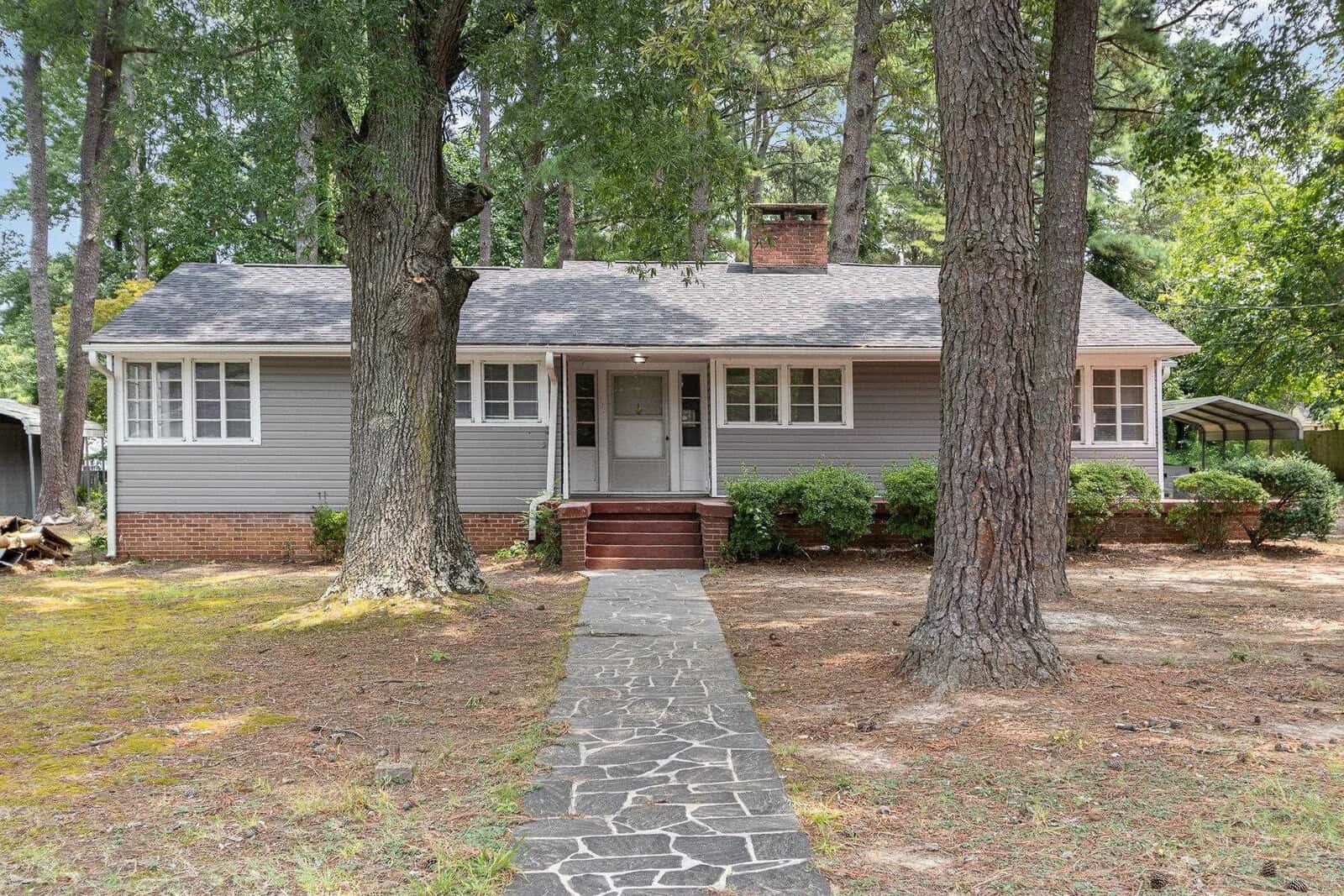 A single-story home with gray siding, featuring a gabled entrance and a brick stoop, set back from a footpath lined with irregular stone slabs and surrounded by mature trees.