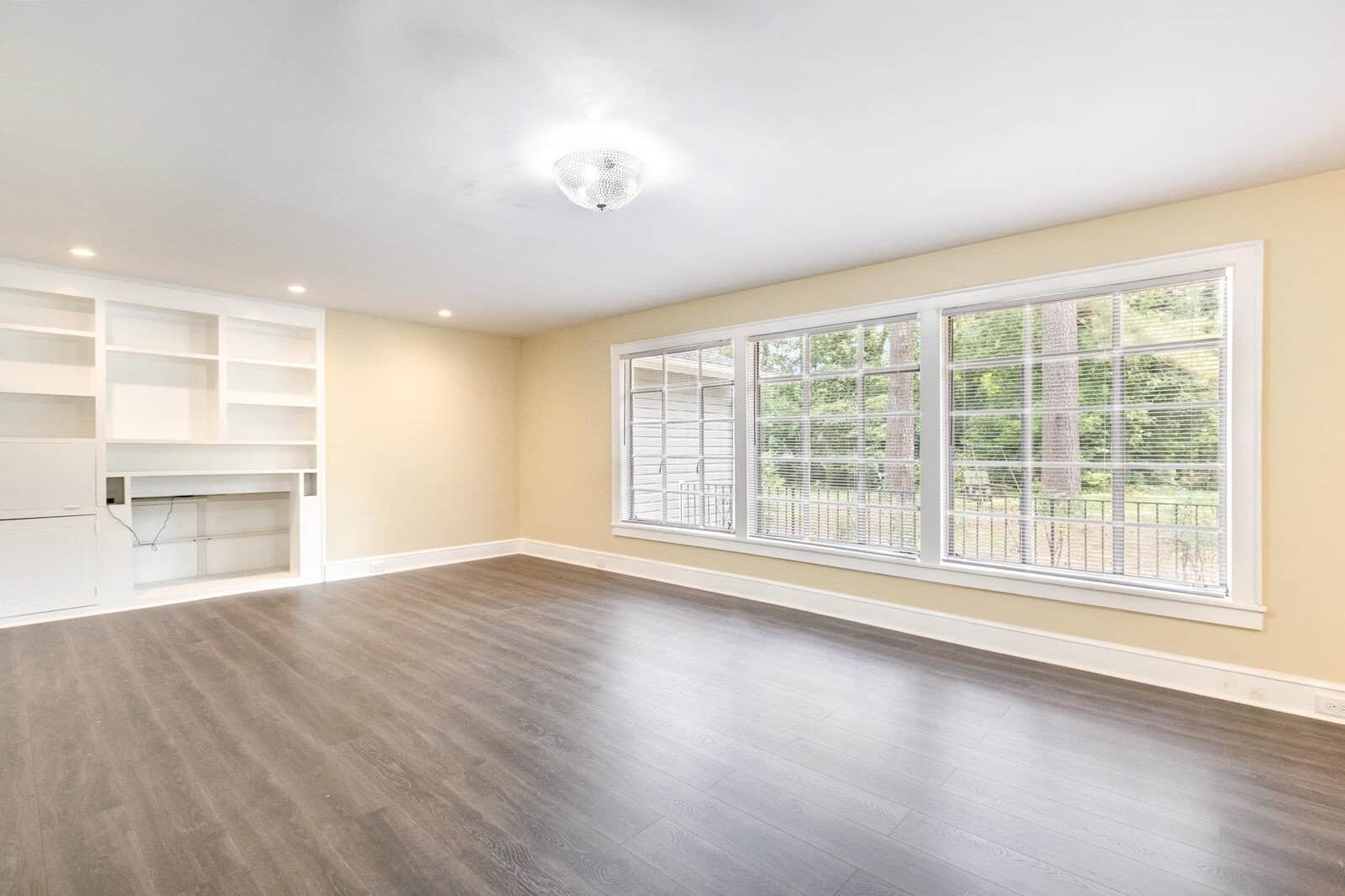 An empty room with light grey flooring, white walls with built-in shelves and cabinets on the left, and a large window with blinds overlooking a fenced outdoor area.