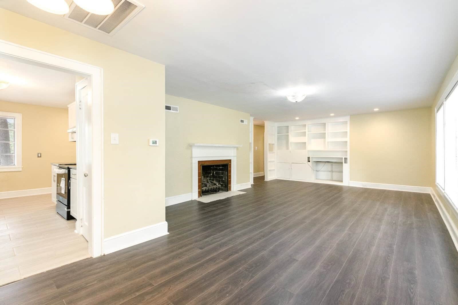 A spacious, empty room with dark wood flooring, a white fireplace, built-in shelving, and an adjacent kitchen area visible through an open doorway.