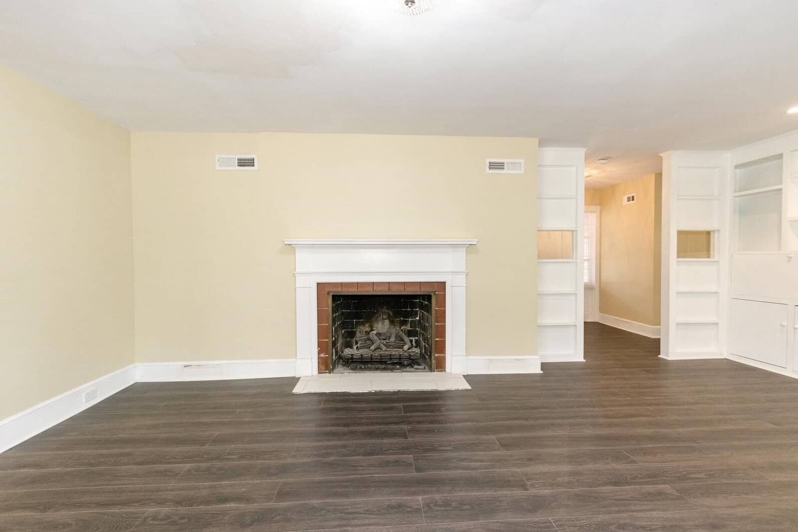 An empty, well-lit room featuring a dark hardwood floor, a white fireplace in the center of the wall, built-in white shelving units to the right, and an open doorway to another room on the left.