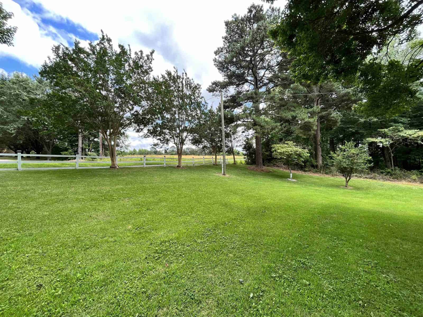 A well-manicured green lawn with a variety of trees under a cloudy sky, enclosed by a white fence.