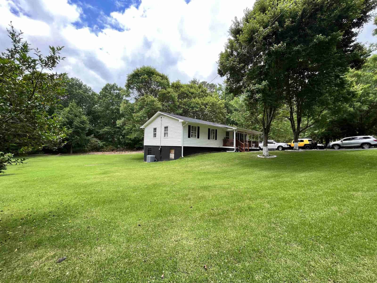 A single-story white house with a dark roof featuring an attached covered porch, surrounded by a well-kept lawn with several trees, under a partly cloudy sky. multiple cars are parked to the side of the house.