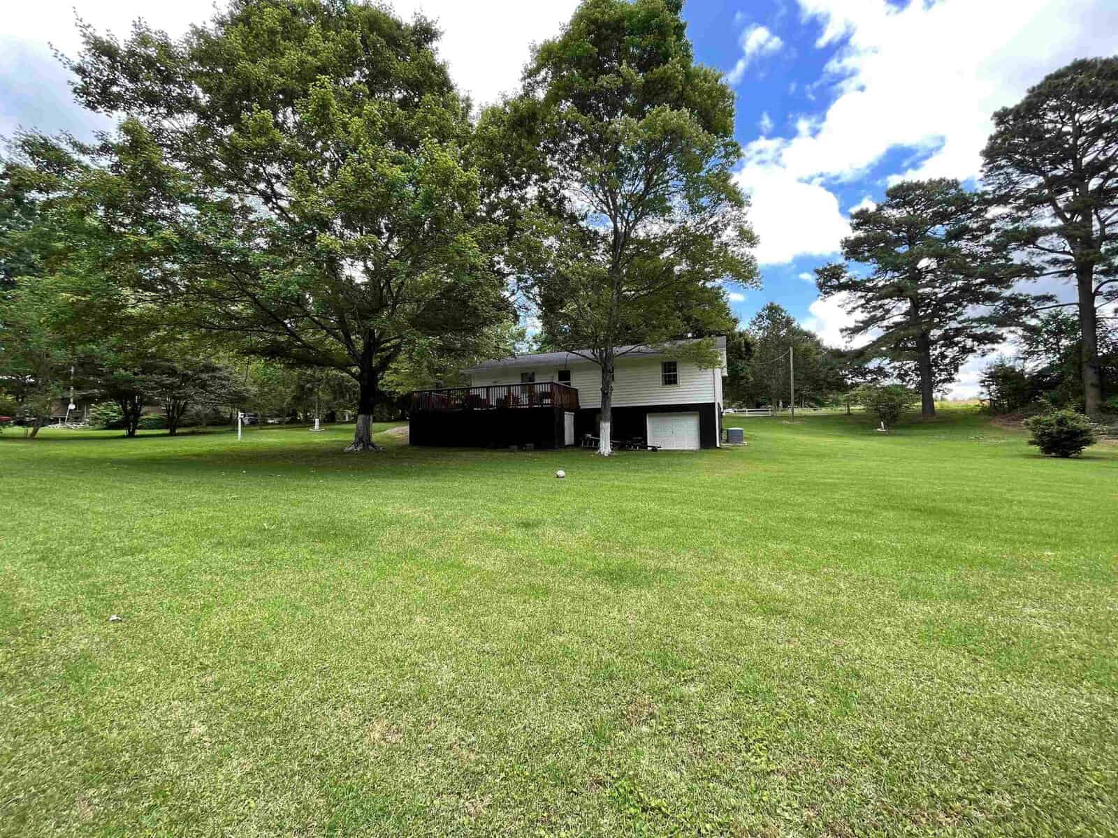 A single-story white house with a black roof and an attached covered porch set in a spacious lawn with several trees under a blue sky with scattered clouds.