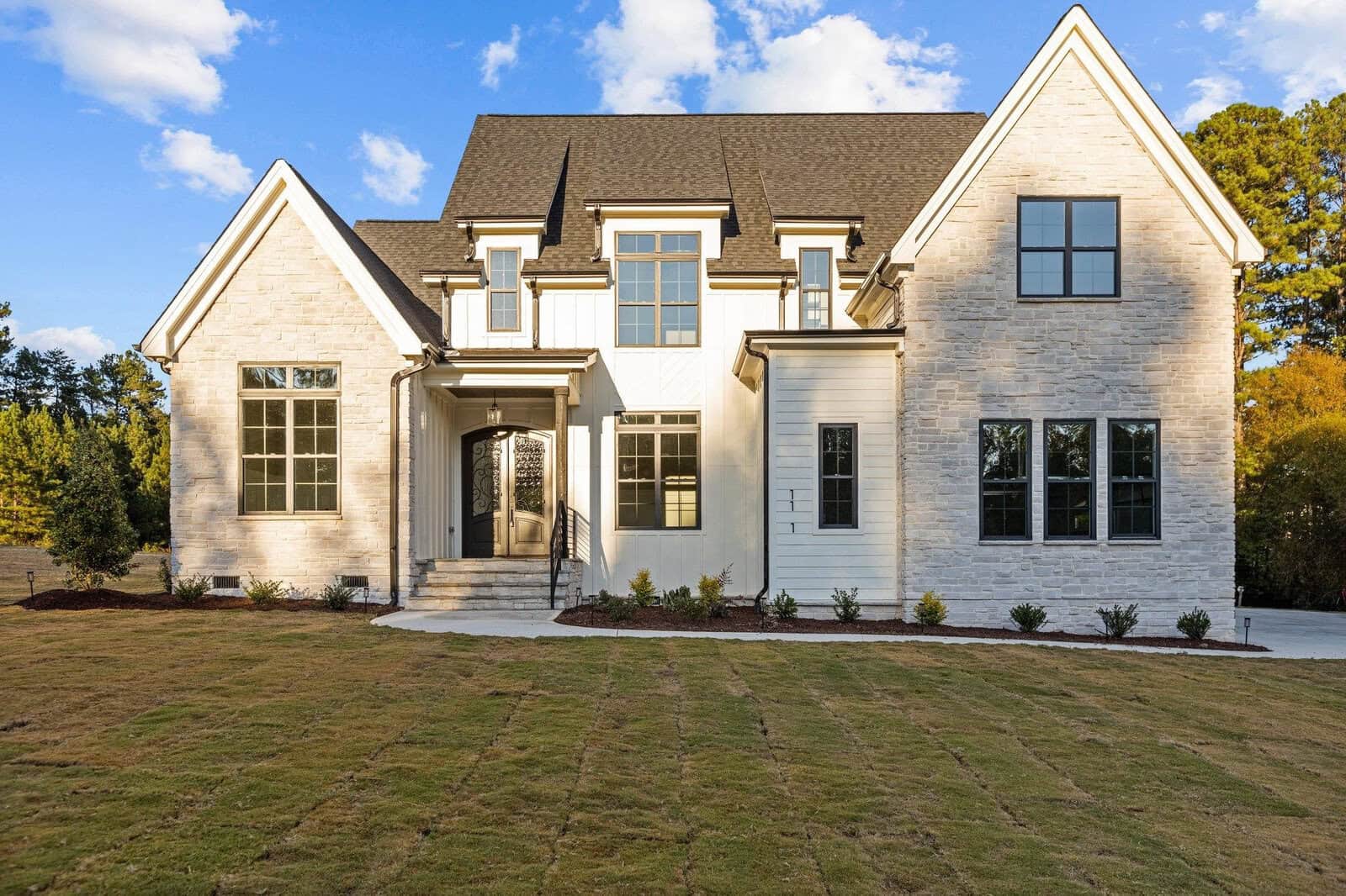 Nestled in the serene surroundings of Wake Forest, this two-story modern house at 1121 Springdale Drive features a light stone facade, dark roof, and several large windows. A well-kept lawn and paved driveway enhance its charm, all under a clear blue sky with a few clouds floating by.