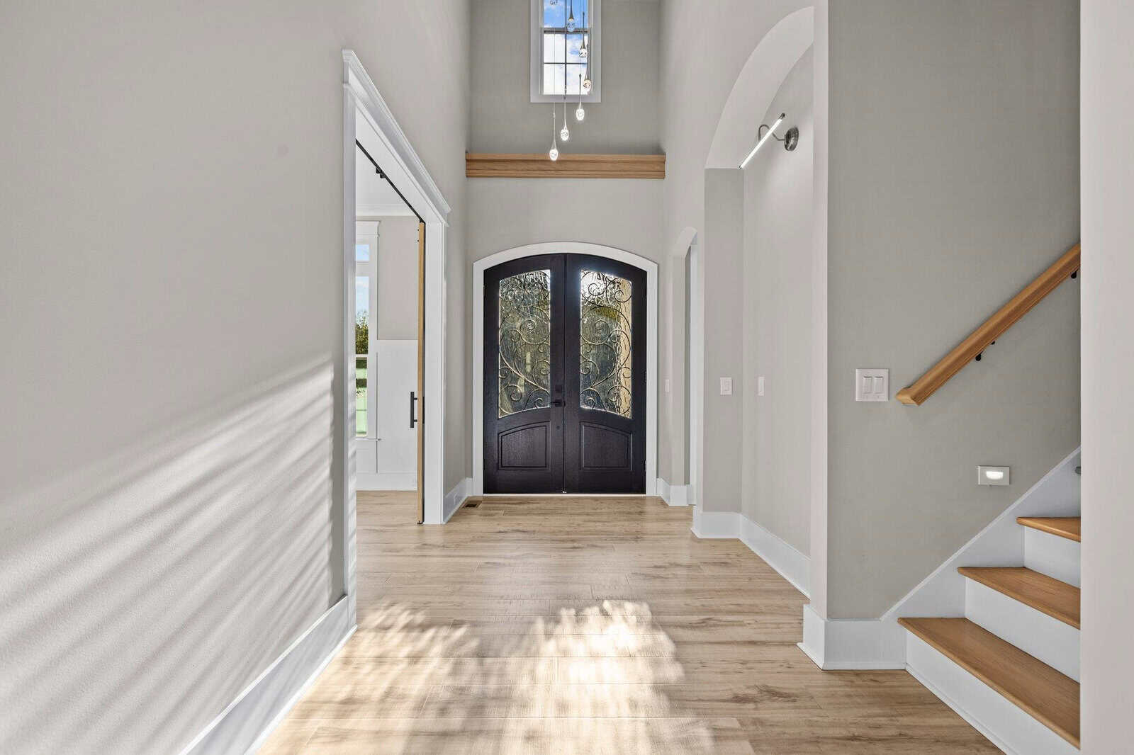 Bright entrance hallway with light wood flooring, arched black doors with ornate glass panels, and a staircase to the right. A large window above the door lets in natural light, creating shadows on the floor. Walls are painted light gray.