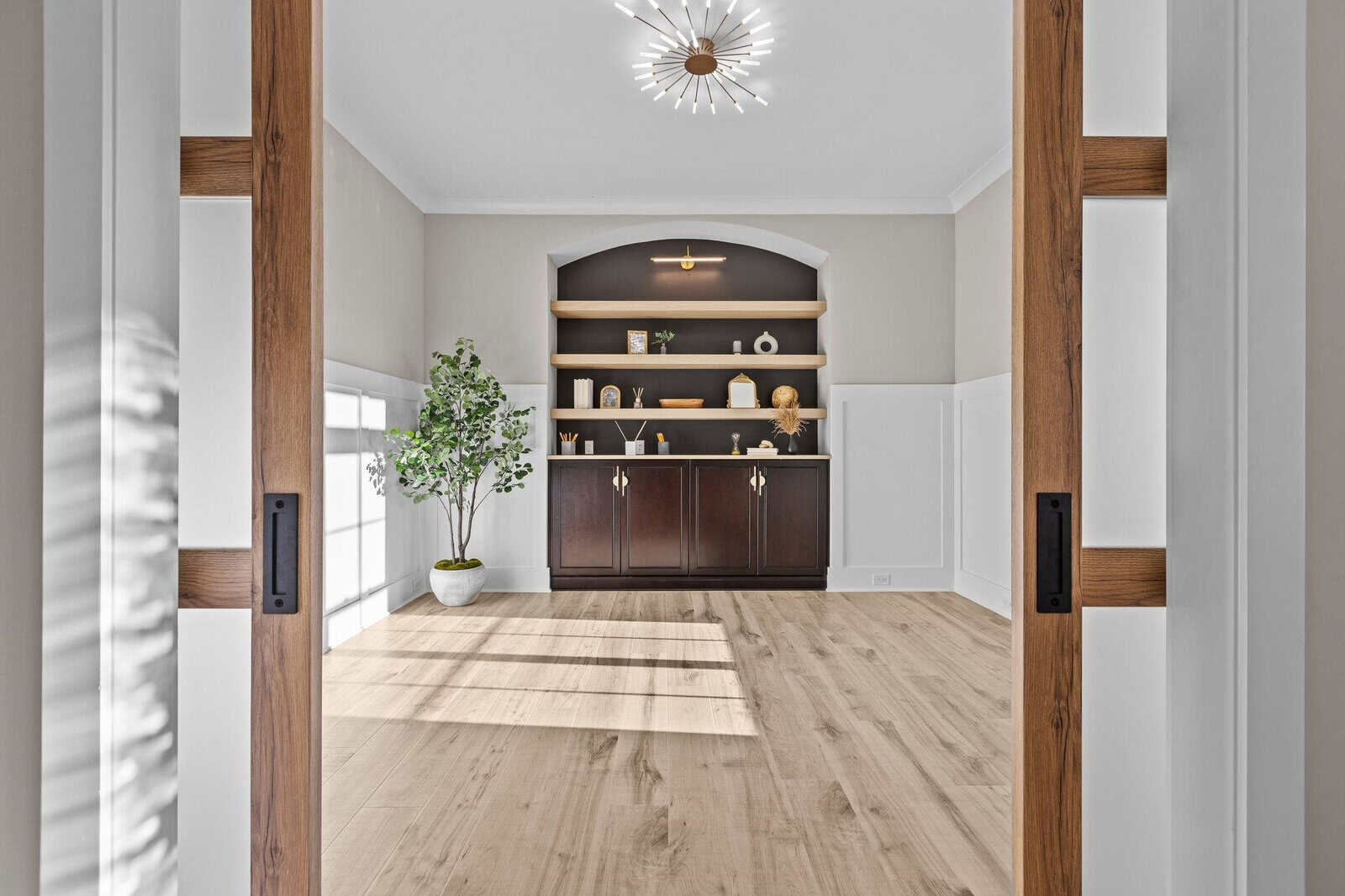 A minimalist room features a wooden floor, a dark wood cabinet with decor items, and built-in shelves. A potted plant sits to the left, and a modern light fixture hangs from the ceiling. Light streams through large windows, creating a warm atmosphere.