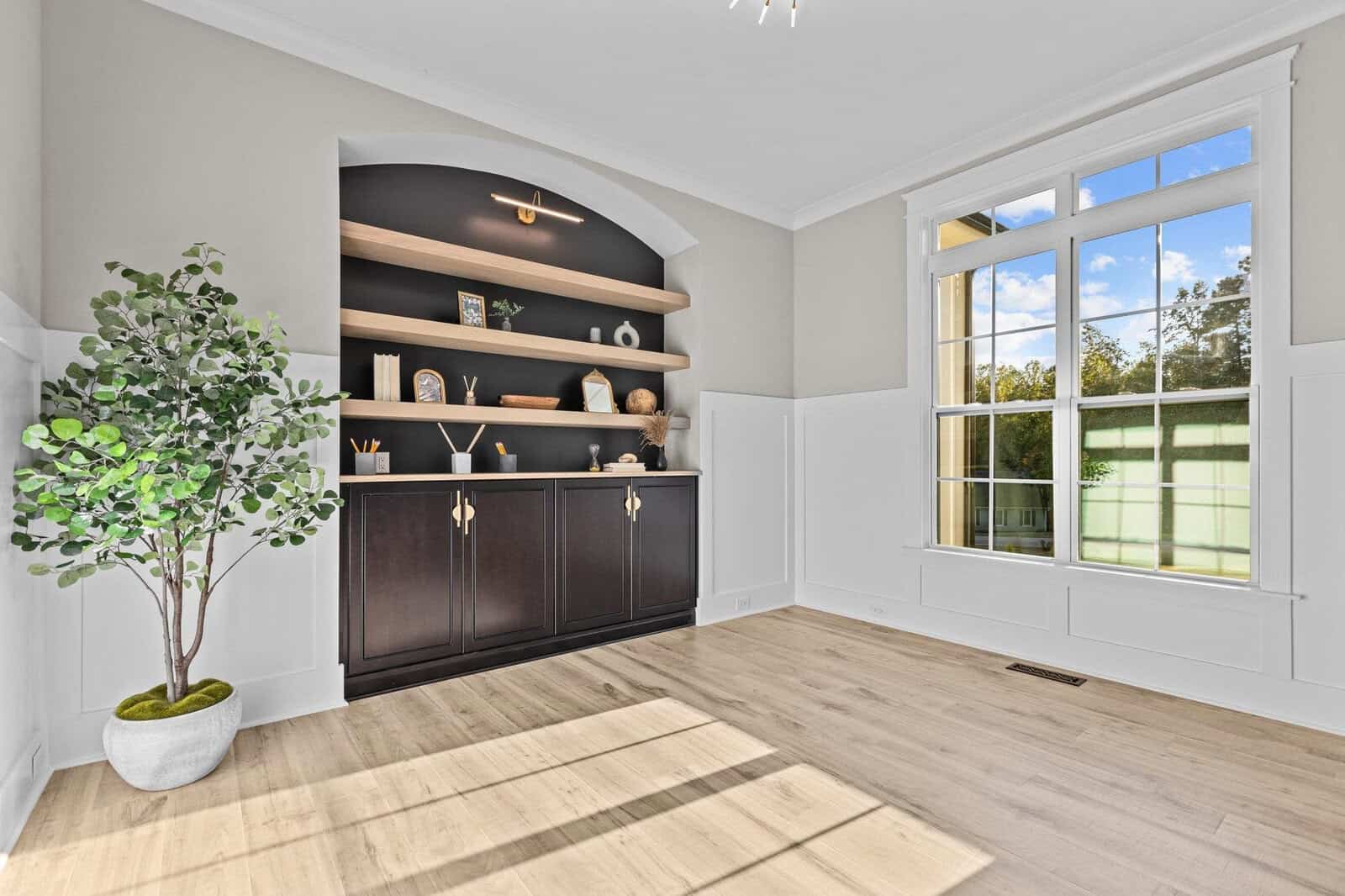 A bright room featuring a large window with a view of trees outside. Inside, there is a black cabinet with open wooden shelves displaying decorative items. A potted plant is placed on light hardwood flooring, bathed in natural light.