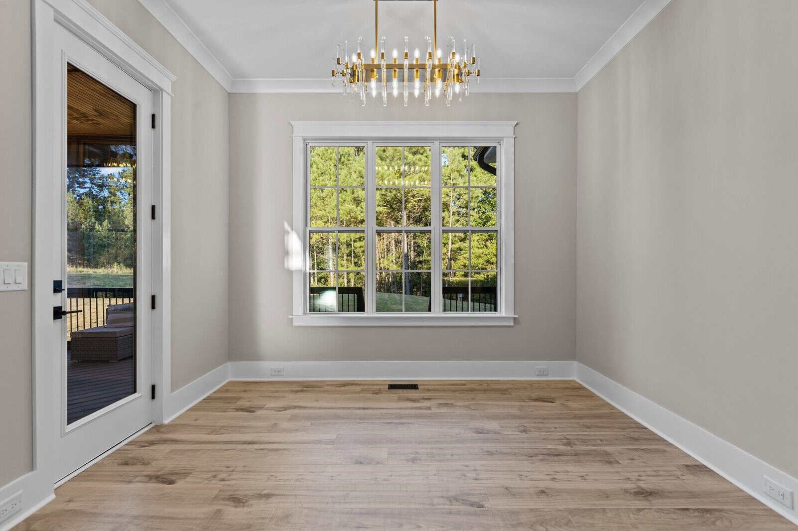A bright, empty room with light wooden floors, grey walls, and a large window showcasing a scenic outdoor view. A modern chandelier hangs from the ceiling, and there's a glass door leading to a patio.
