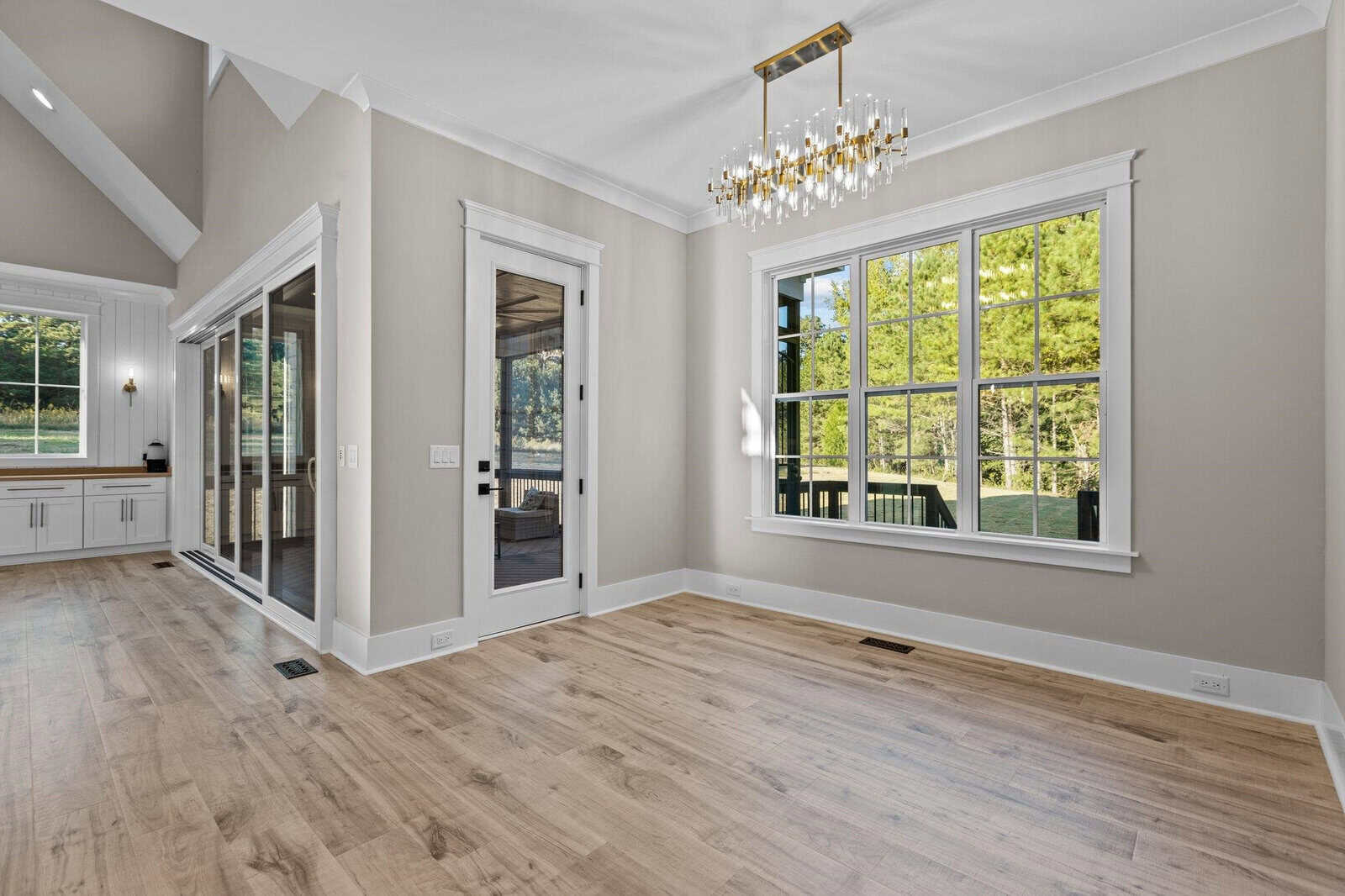 A bright, empty room with light wood flooring, large windows, and a glass door leading outside. A modern chandelier hangs from the ceiling. Adjacent is a sliding glass door with a view of trees. A kitchen area is partially visible in the background.