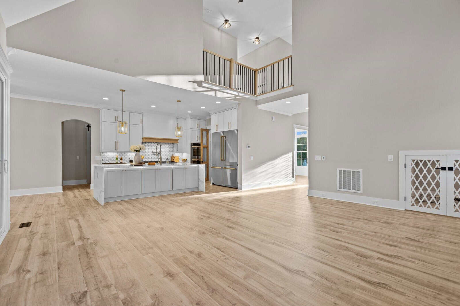 A spacious, modern open-plan kitchen and living area with light wood flooring. The kitchen features white cabinets, a large island, and stainless steel appliances. Above, a high ceiling with recessed lights and a wooden railing overlooks the space.