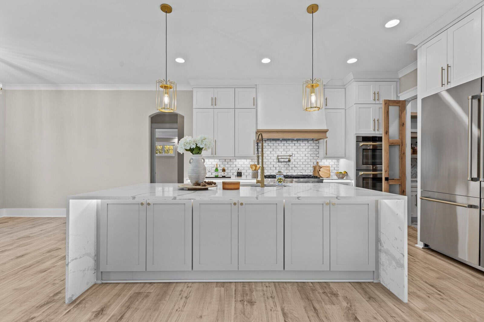 A modern kitchen with a large white marble island, pendant lights, and white cabinets. Stainless steel appliances, including a refrigerator and double oven, are visible. The backsplash features white subway tiles. A vase with flowers sits on the counter.