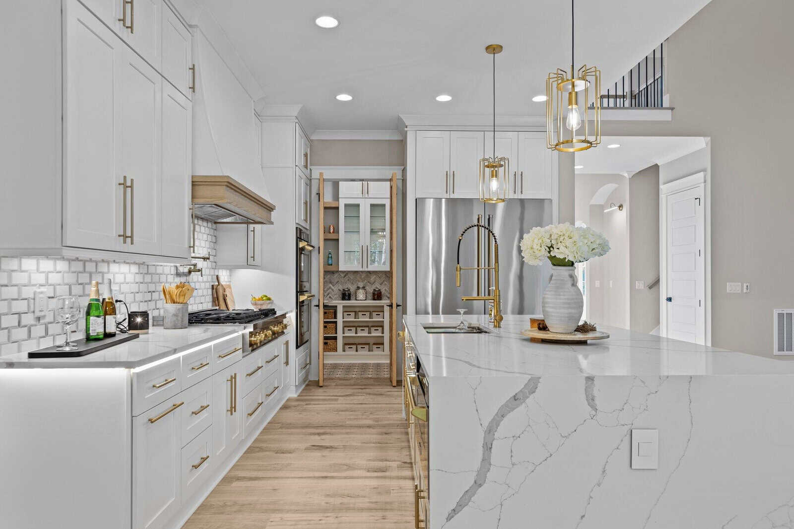 Modern kitchen with white cabinets, a large marble island, and stainless steel appliances. The space features pendant lights, a backsplash with patterned tiles, and a wine rack in the background. A vase with flowers decorates the island.