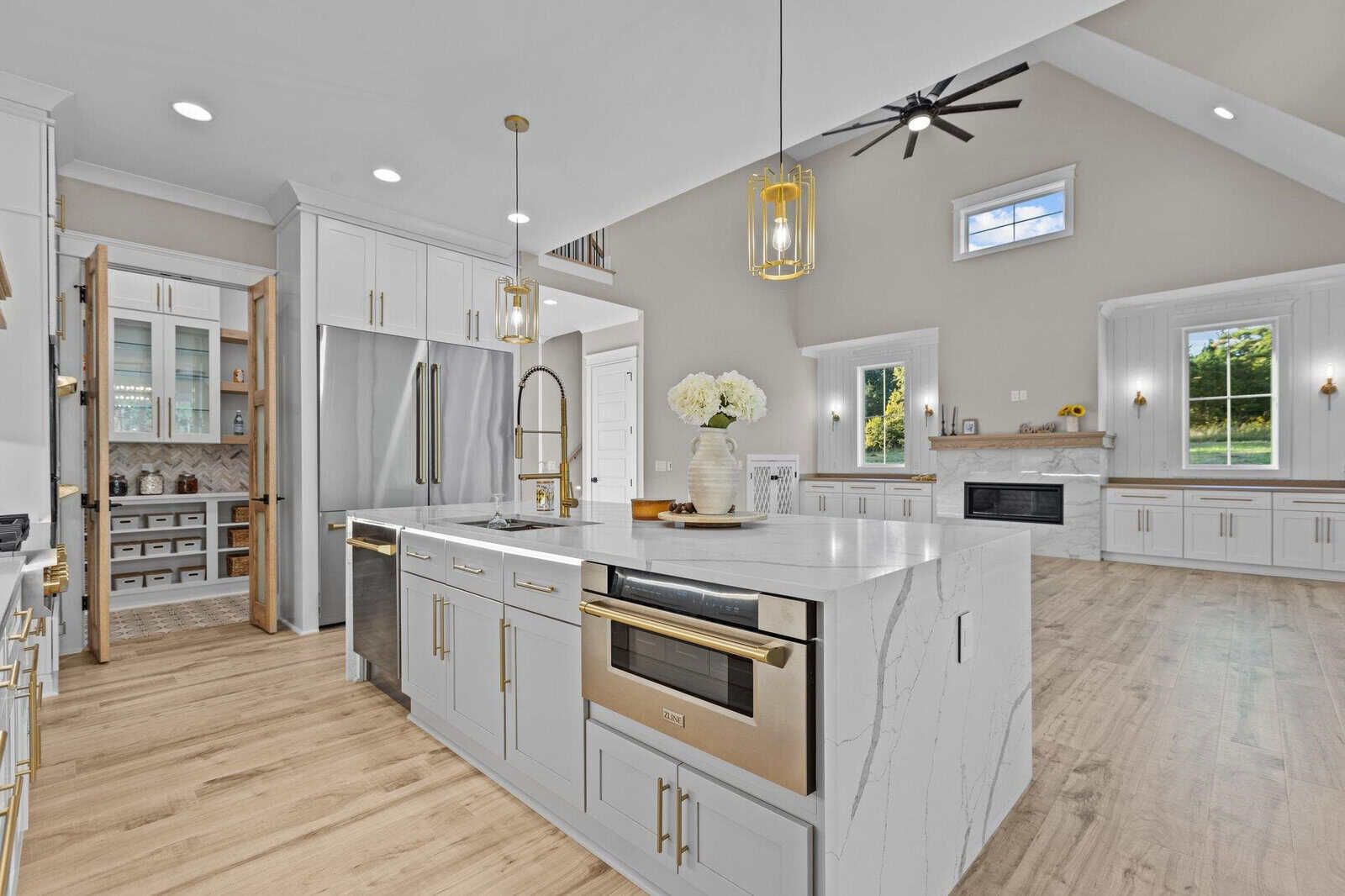 Modern kitchen with white cabinetry, marble countertops, and hardwood flooring. Features include a large island with a built-in microwave, double-door fridge, and pendant lights. A pantry with shelving is visible, along with a fireplace in the adjoining room.