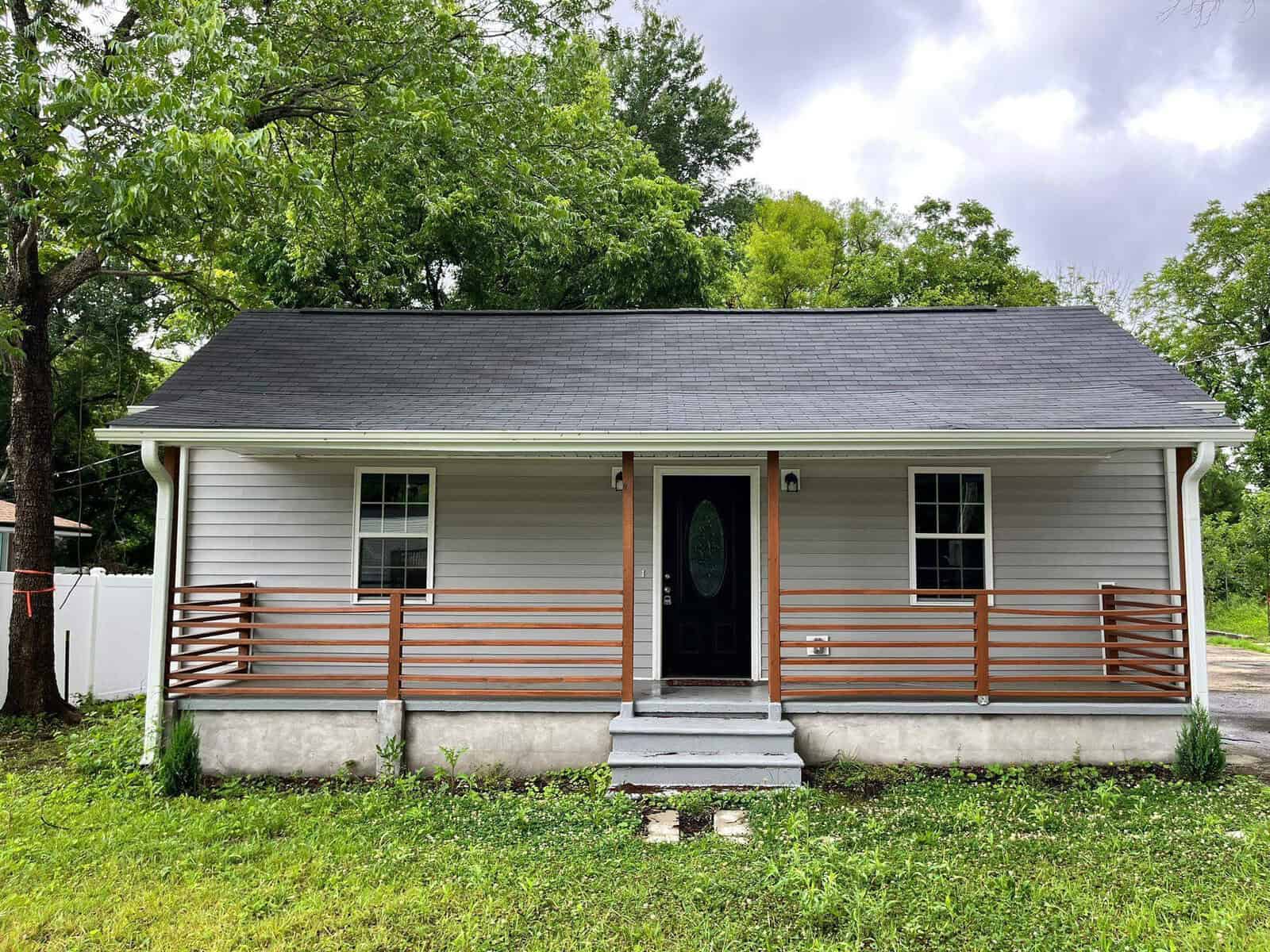 A single-story house with white siding, a gray shingle roof, and a front porch with wooden railings. the yard is green with grass and surrounded by trees.
