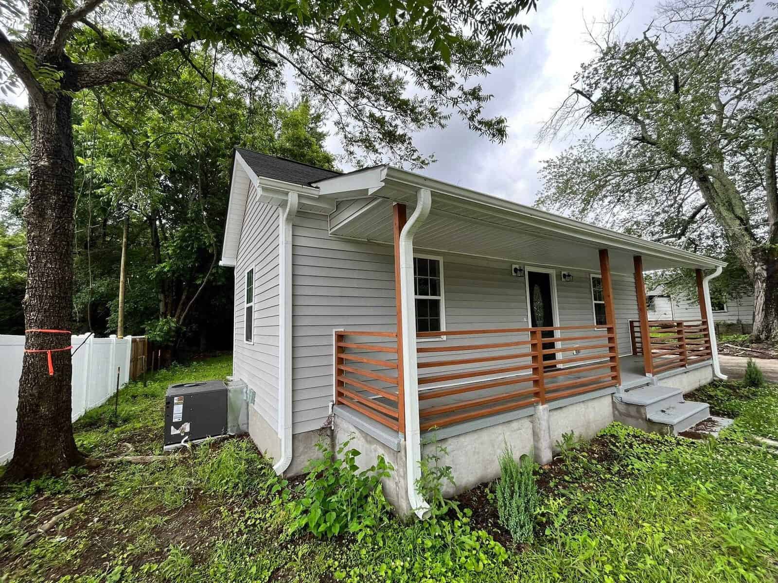 A small single-story house with gray siding and a wooden porch railing, situated among trees with an overcast sky above.