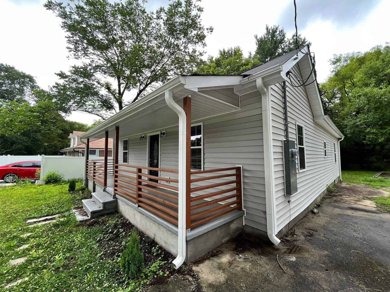A single-story house with white siding, featuring a front porch with a slatted wooden railing, surrounded by greenery, with a red vehicle visible in the background.