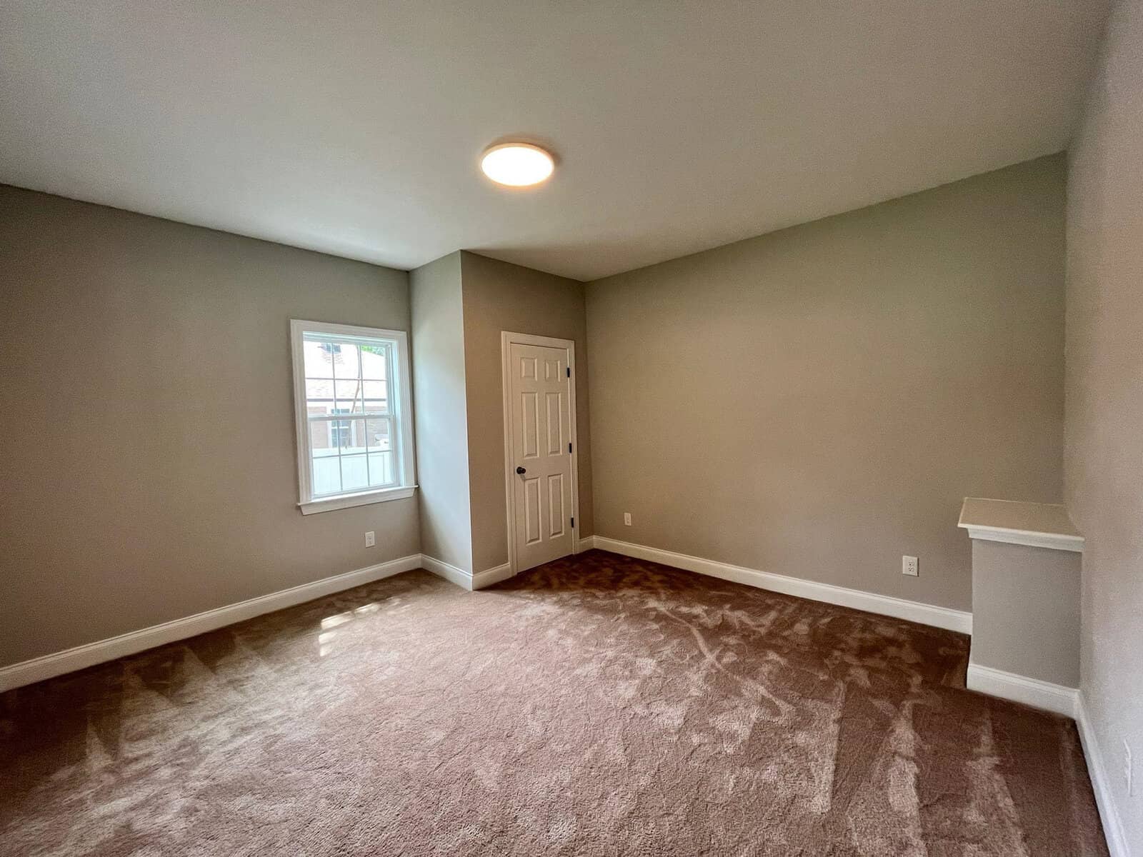 An empty room with beige carpeting, light gray walls, a window on the left wall, and a closed white door on the far wall.