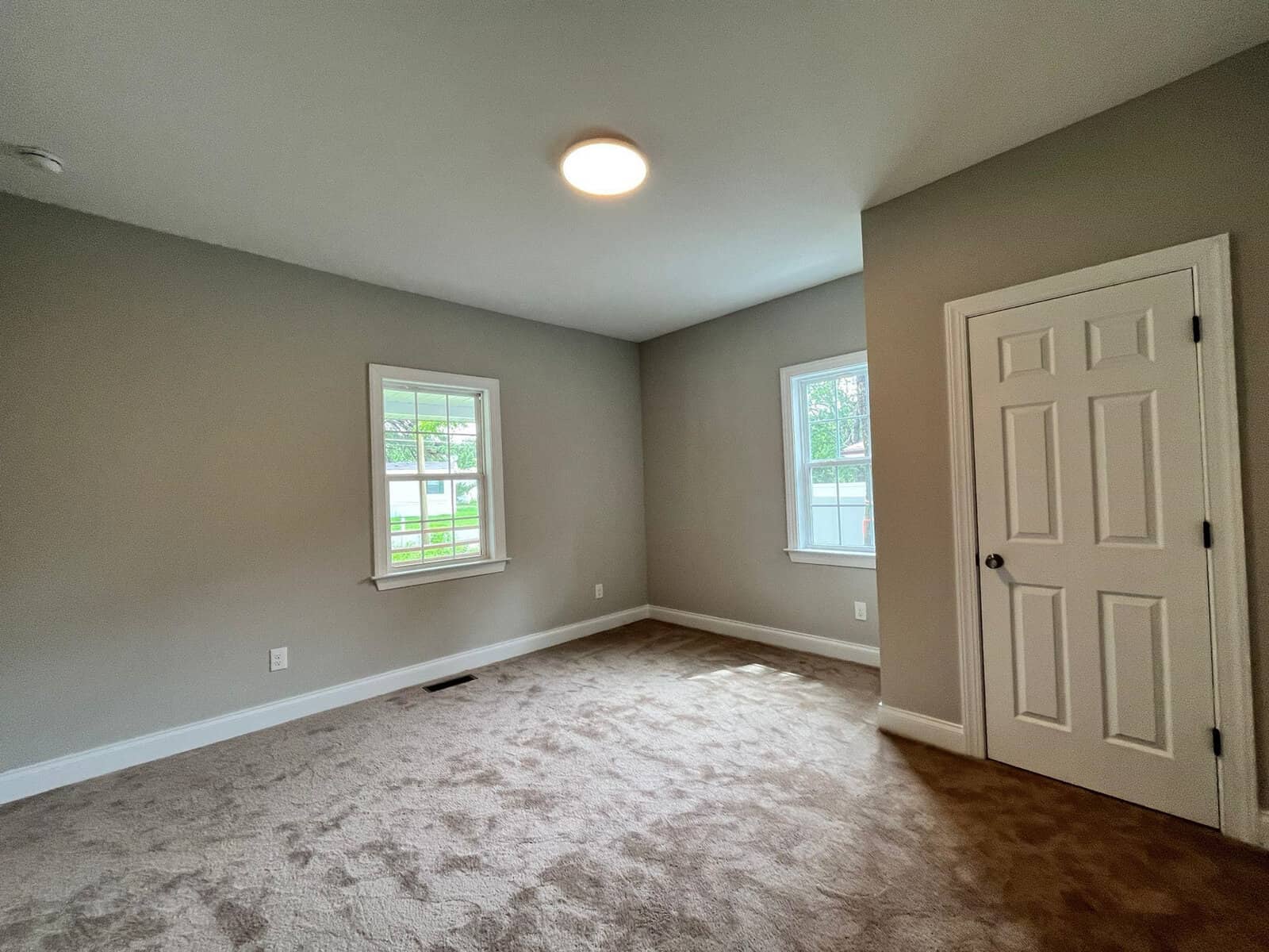 An empty room with light gray walls, beige carpet flooring, one window with white trim, and a white closed door on the right.