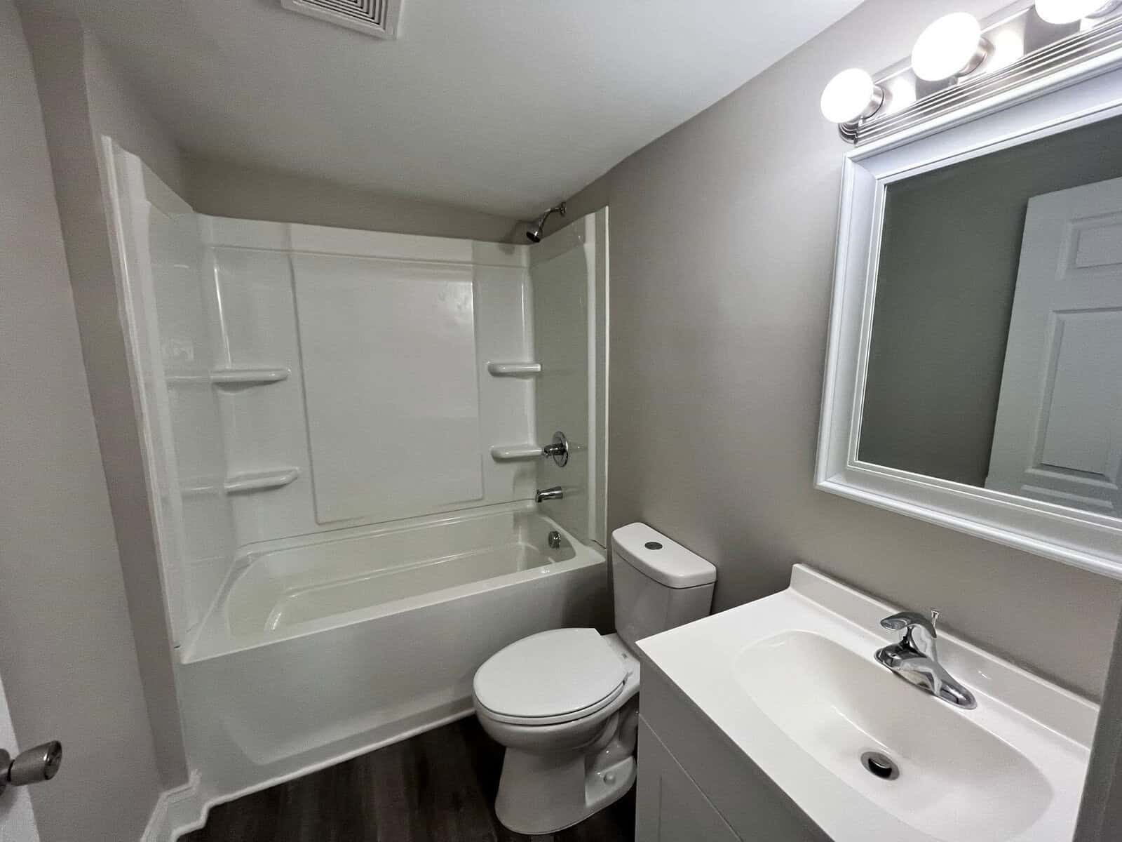 A modern bathroom interior featuring a white bathtub with an overhead shower, a toilet to the left, a vanity with a sink and a mirror with bright lights above it, set against gray walls and a dark floor.