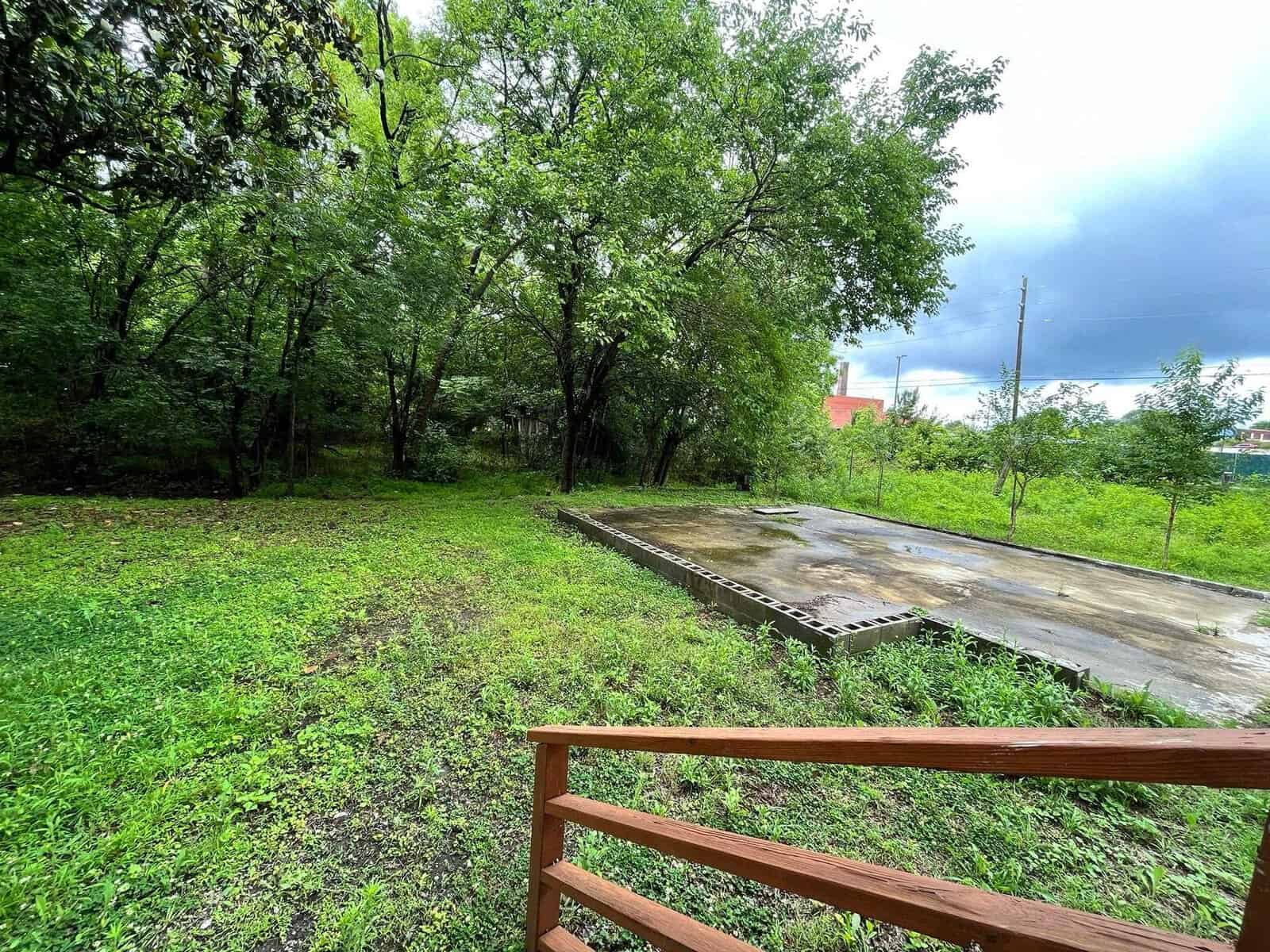 A wooden fence in the foreground overlooks a green area with a mix of grass and trees, and a concrete slab in the midst of the natural foliage, with overcast skies above.
