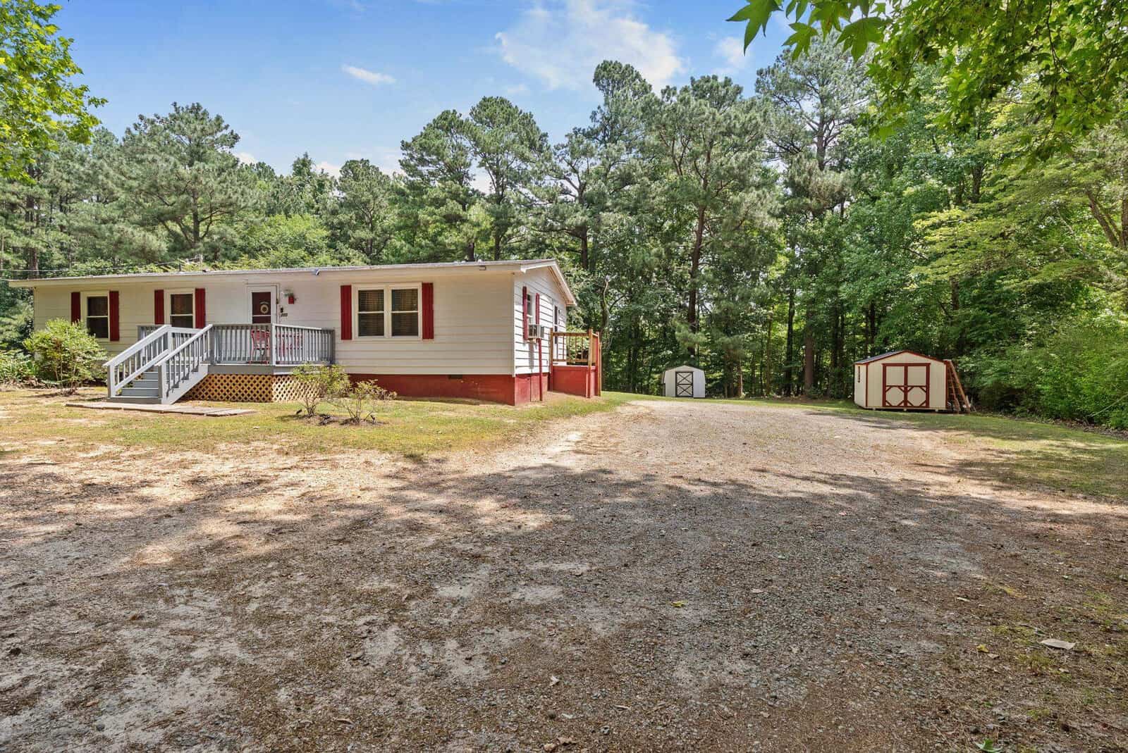 A single-story home with white siding and red trim, featuring a front deck with steps leading up to the entrance, situated on a lot with trees in the background and a small storage shed to the right. there is a gravel area in front of the house that could serve as a driveway.