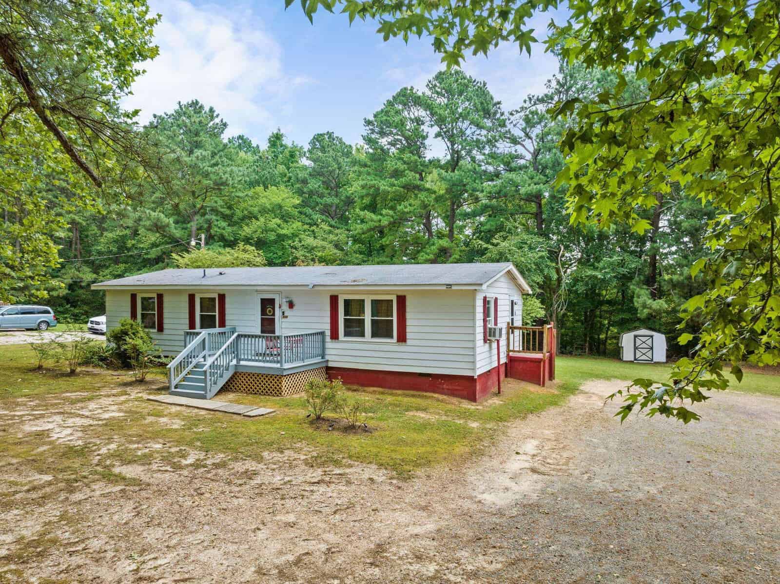 A single-story red and white house with a front porch, surrounded by green trees, with a gravel driveway and a white vehicle parked to the side.