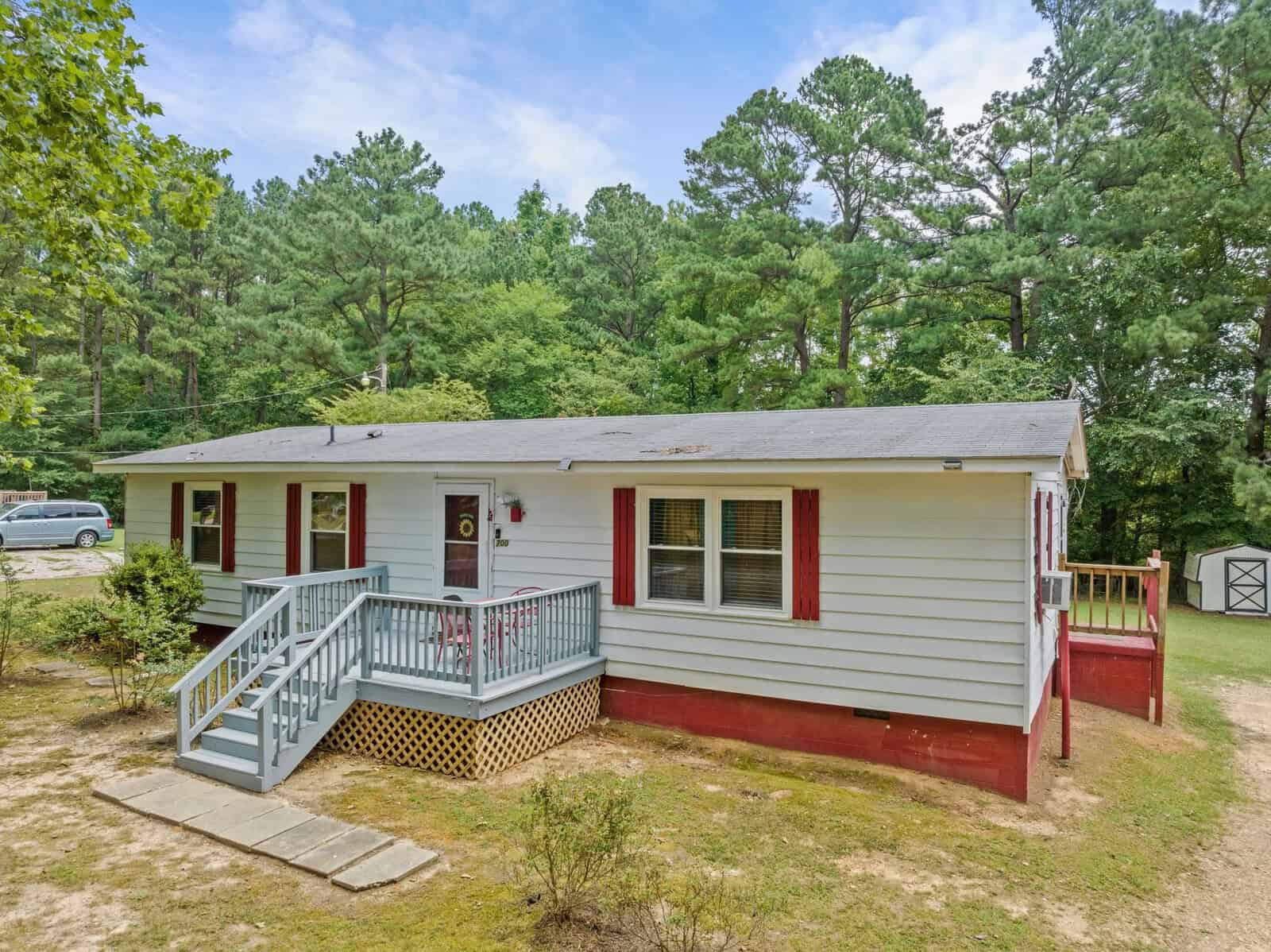 A single-story house with white siding and red accents, featuring a small front porch and an adjacent driveway with a car parked in it, surrounded by trees and a lawn area.