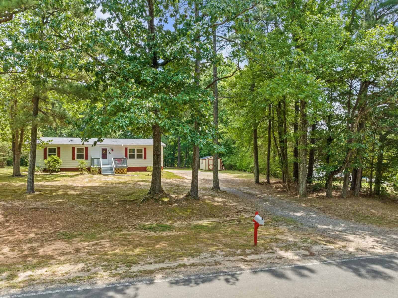 A single-story house with a red and white exterior is nestled among tall trees on a sunny day, with a gravel driveway leading up to it and a red mailbox at the roadside.