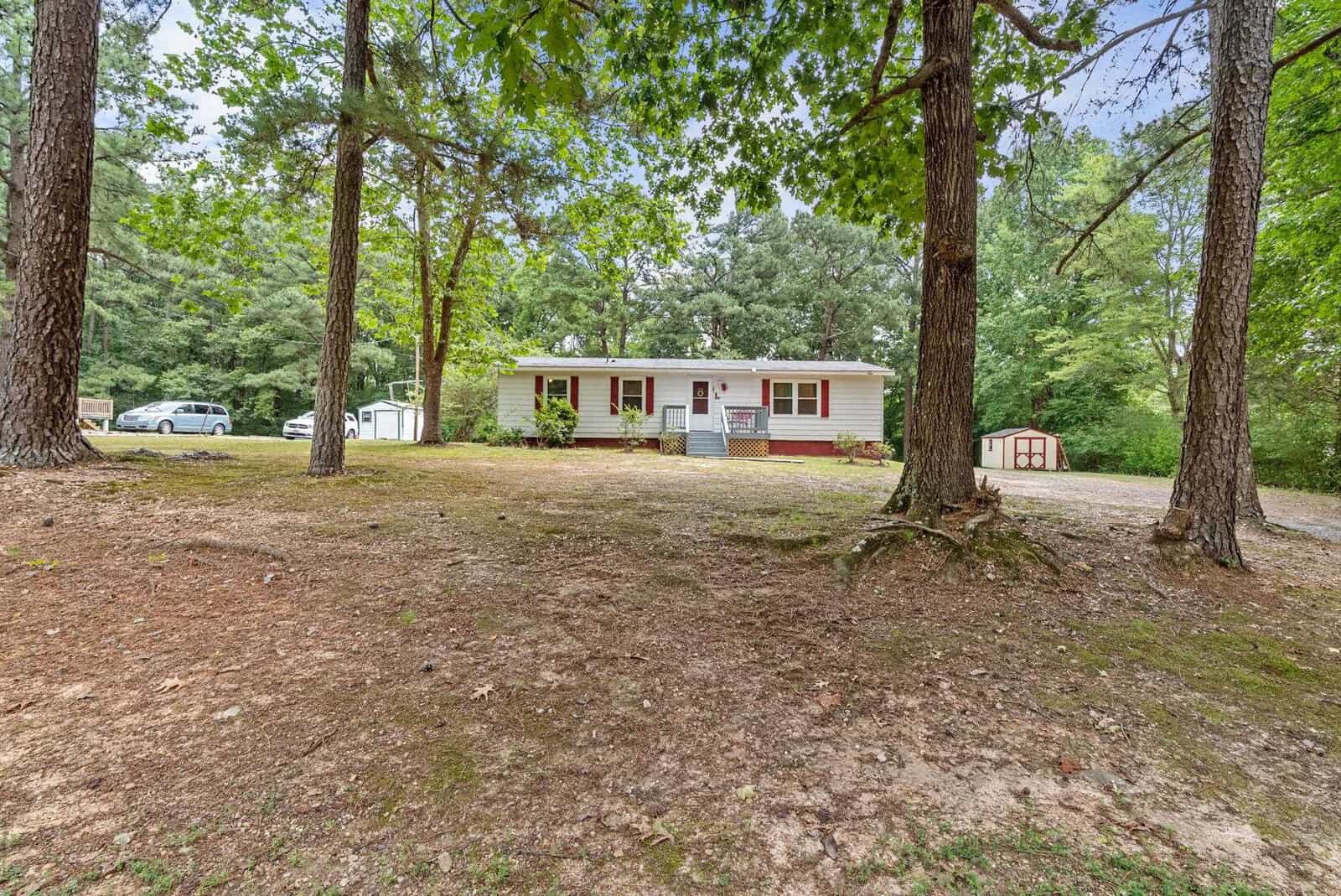 A single-story white house with red shutters and a front porch set among mature trees with a cleared, grassy front yard and a small outbuilding to the right. a car is parked to the left side of the house.